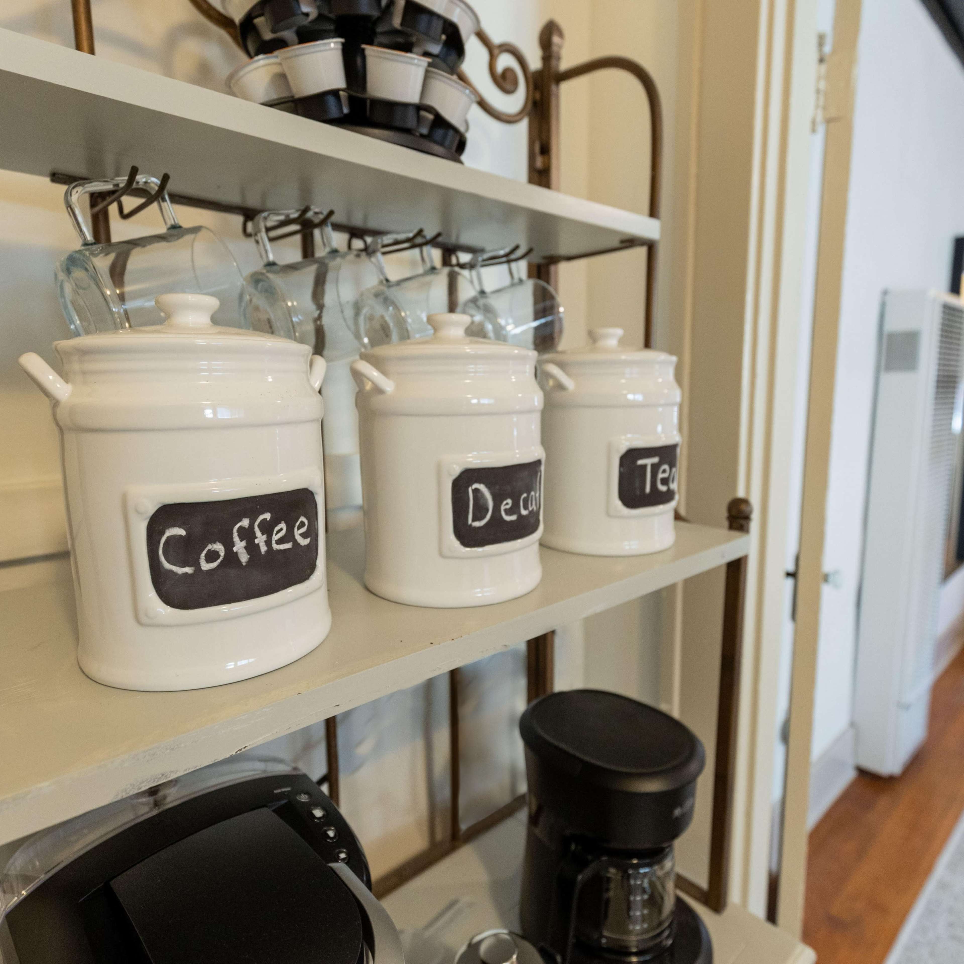 A shelf with labeled white containers for coffee, decaf, and tea, alongside a coffee maker and glassware.
