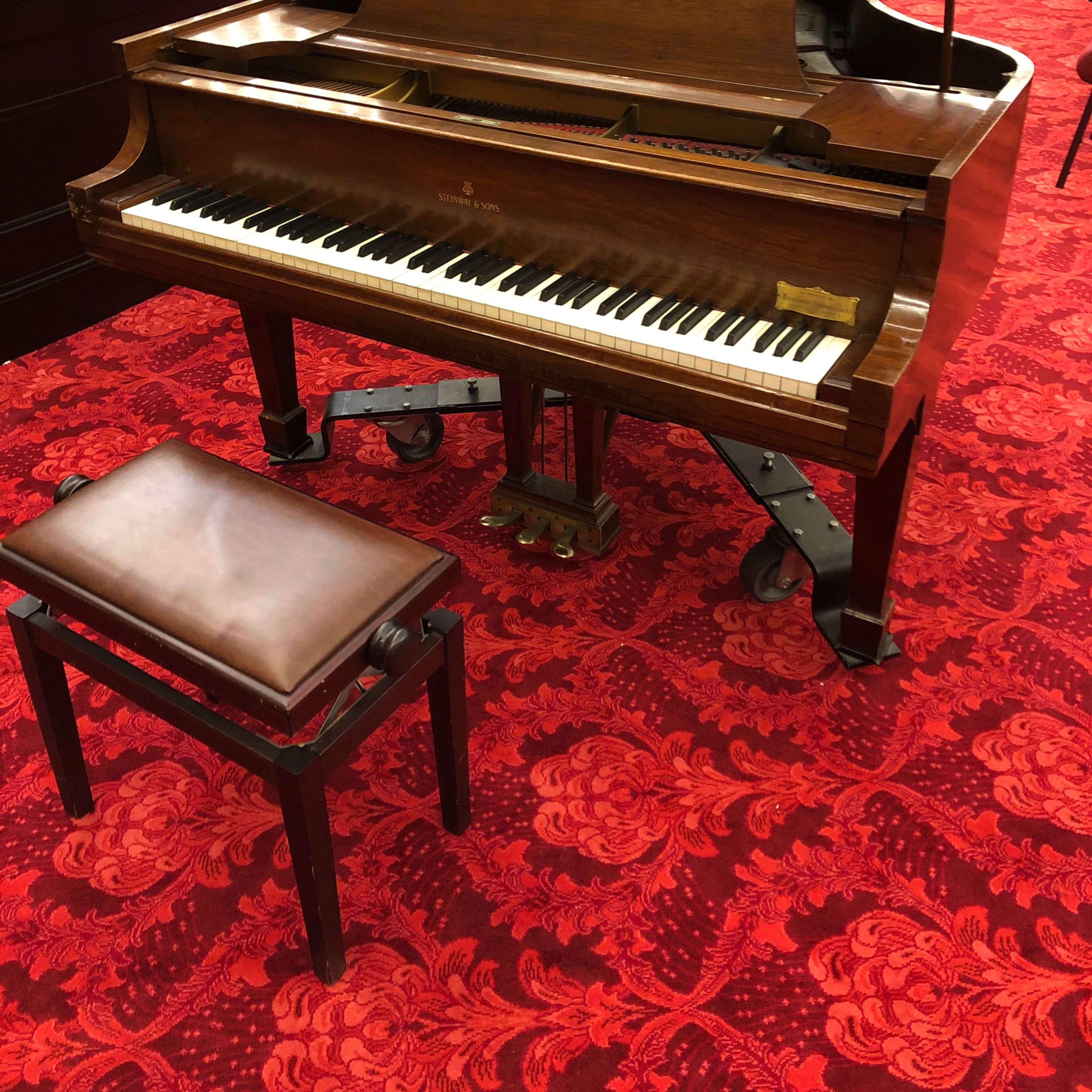 A grand piano with a matching bench is placed on a red carpet in a room with empty chairs arranged nearby.