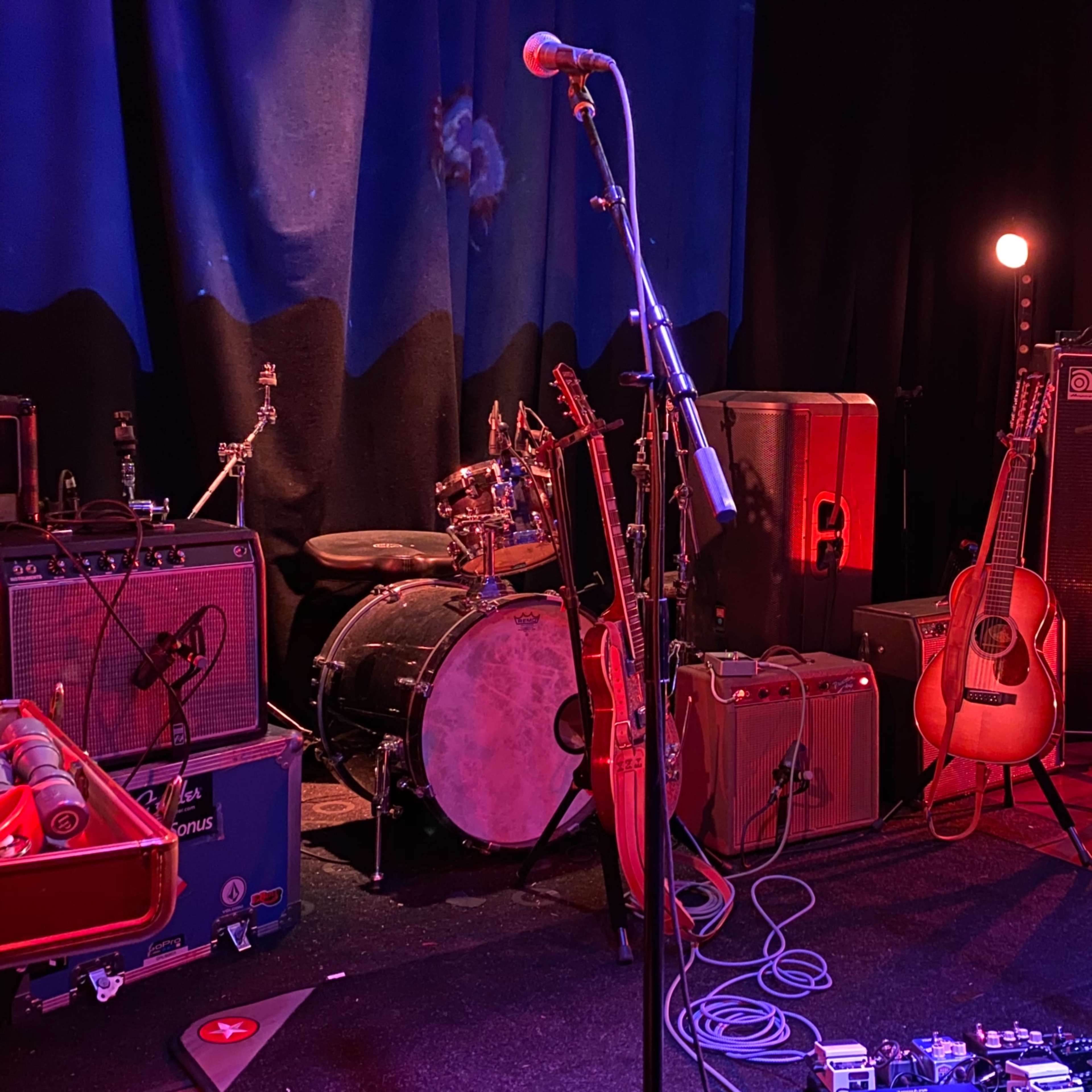 The stage is set with various musical instruments, including guitars, amplifiers, a drum kit, and microphones, ready for performance.