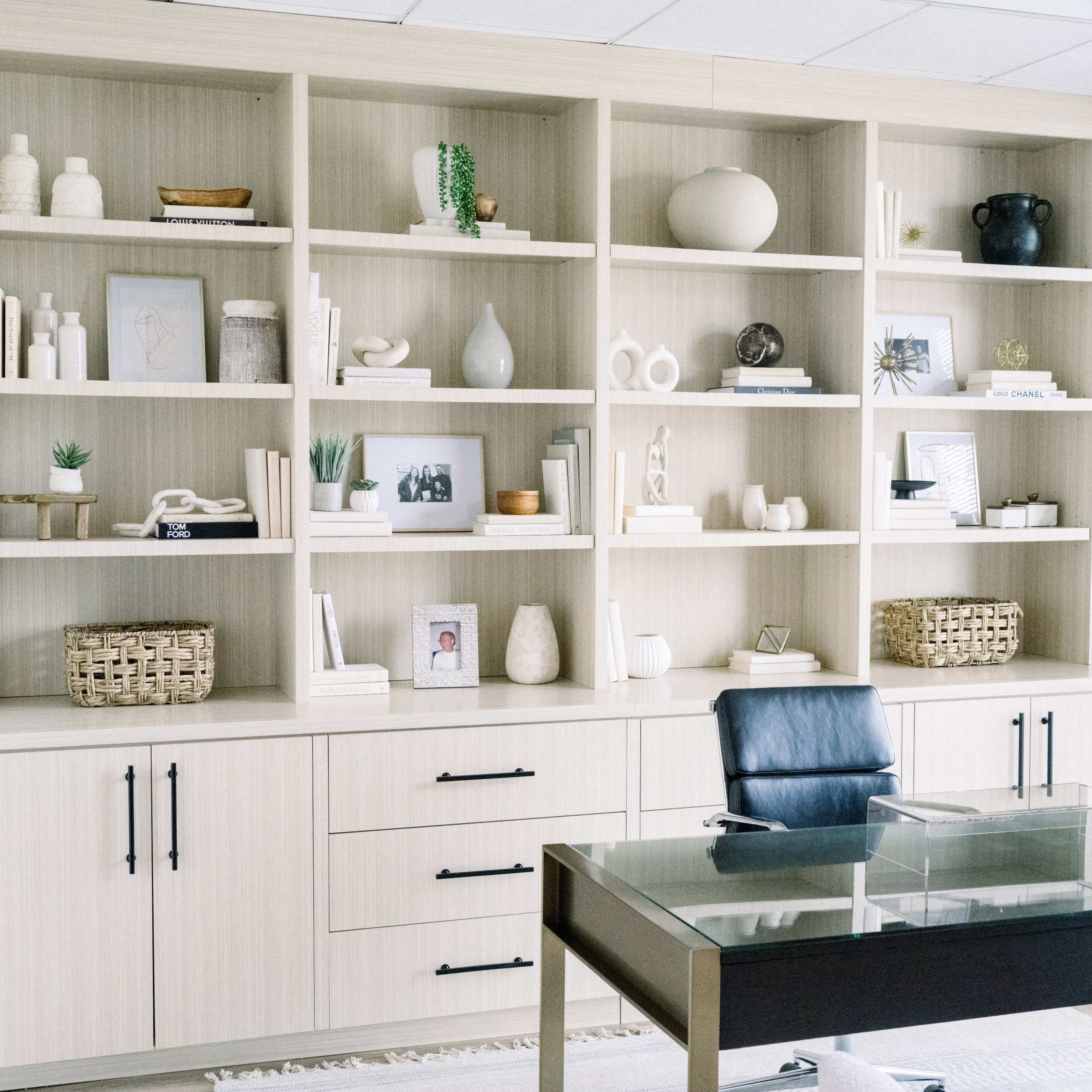 A modern office space features a tall shelving unit filled with decorative items and photographs, alongside a sleek desk and a chair.