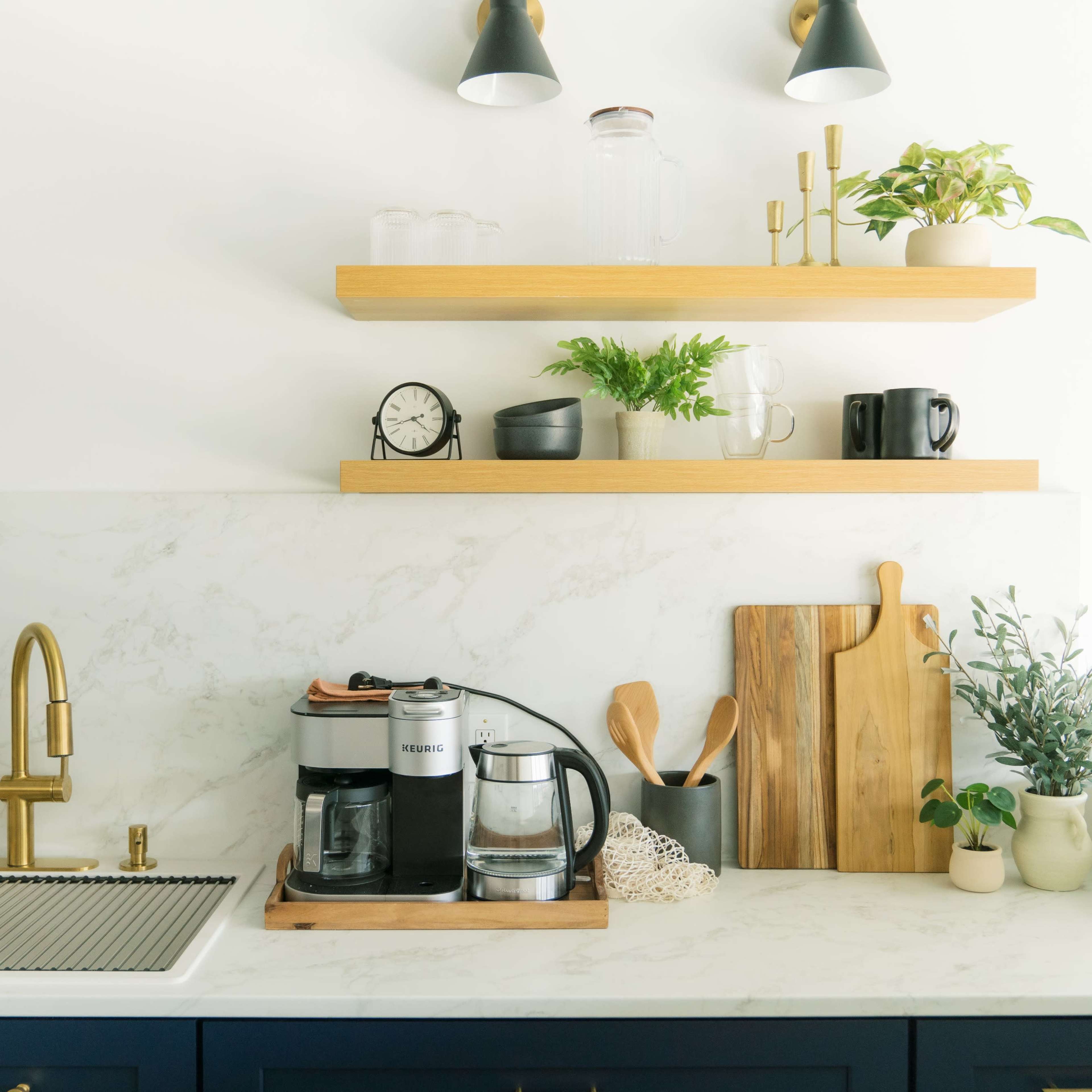 A modern kitchen features a coffee maker on a wooden tray, accompanied by utensils and plants on a floating shelf above a marble countertop.