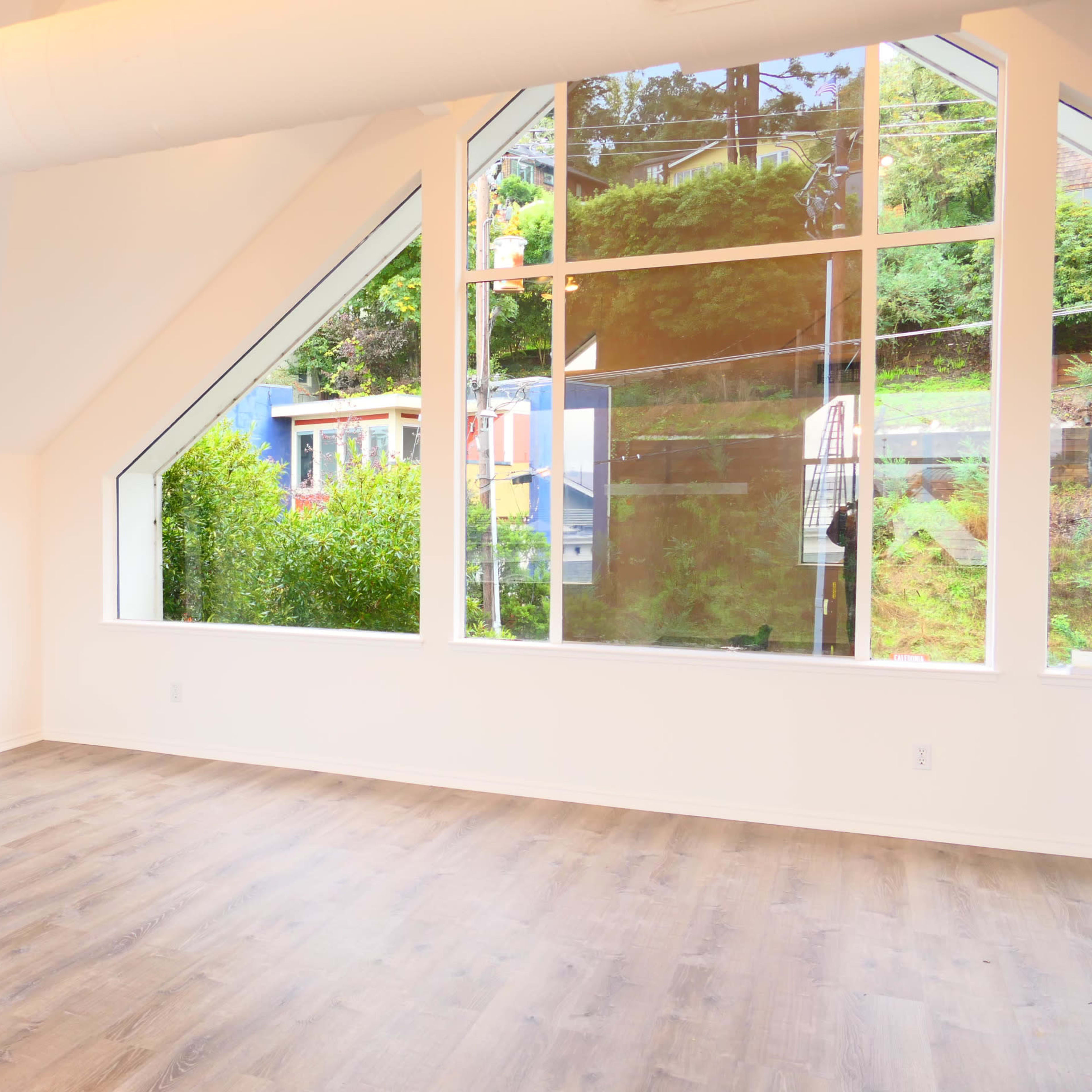 A spacious room with large triangular windows overlooking greenery, and a light-colored wood floor.