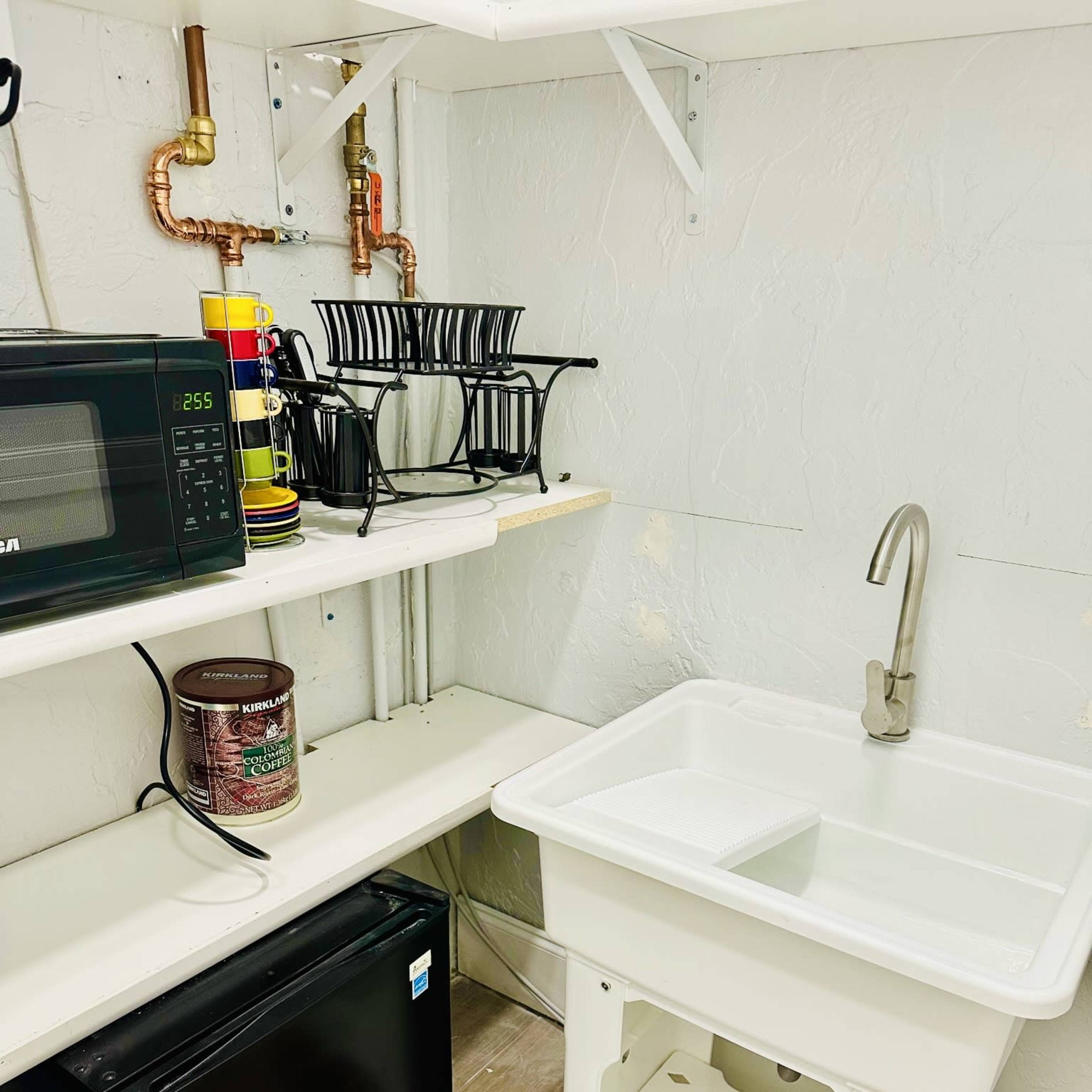 A small utility area featuring a sink, a black refrigerator, a microwave, and a rack for kitchen items.