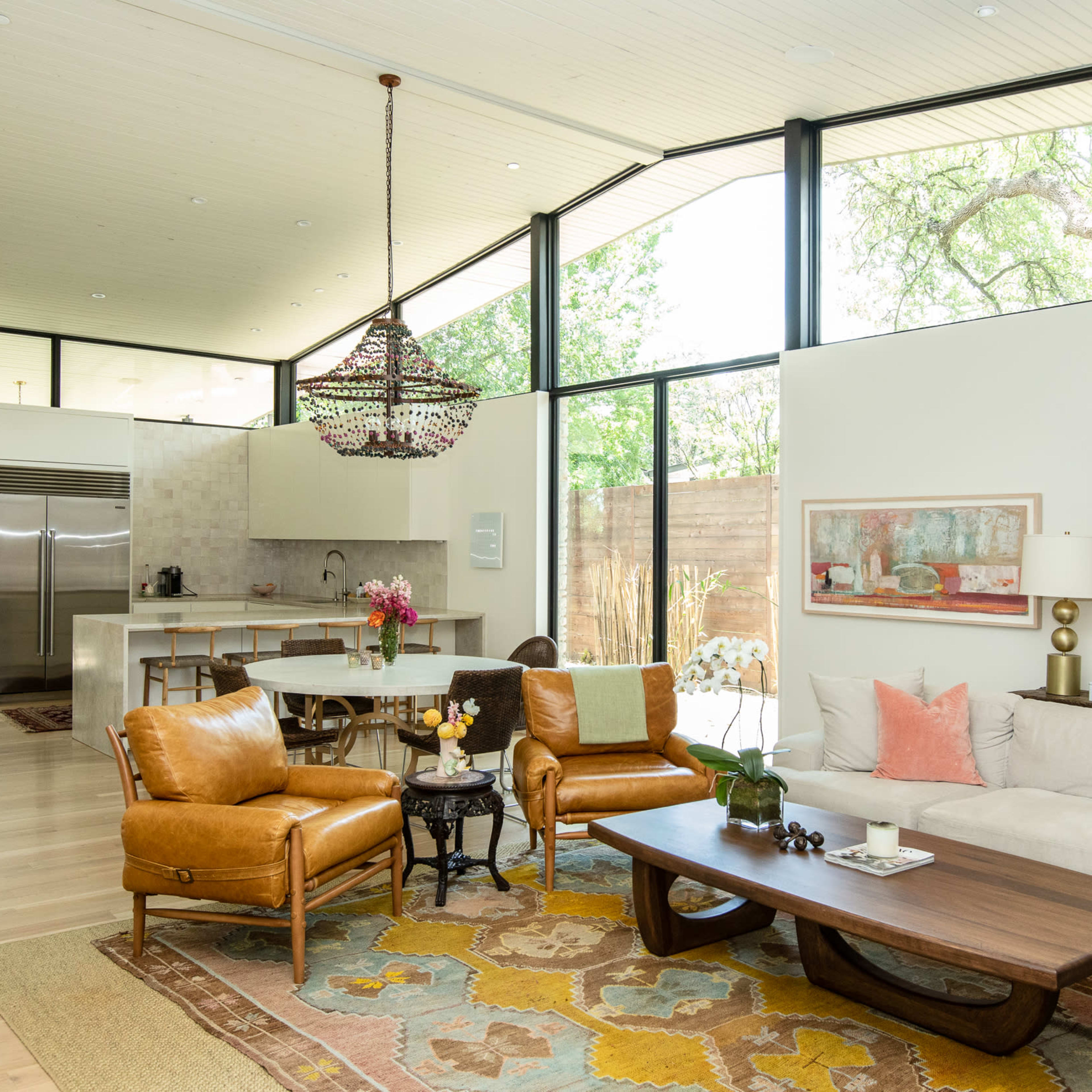 A modern living room with large windows allowing natural light, featuring brown leather chairs, a beige sofa, a wooden coffee table, and a decorative rug.