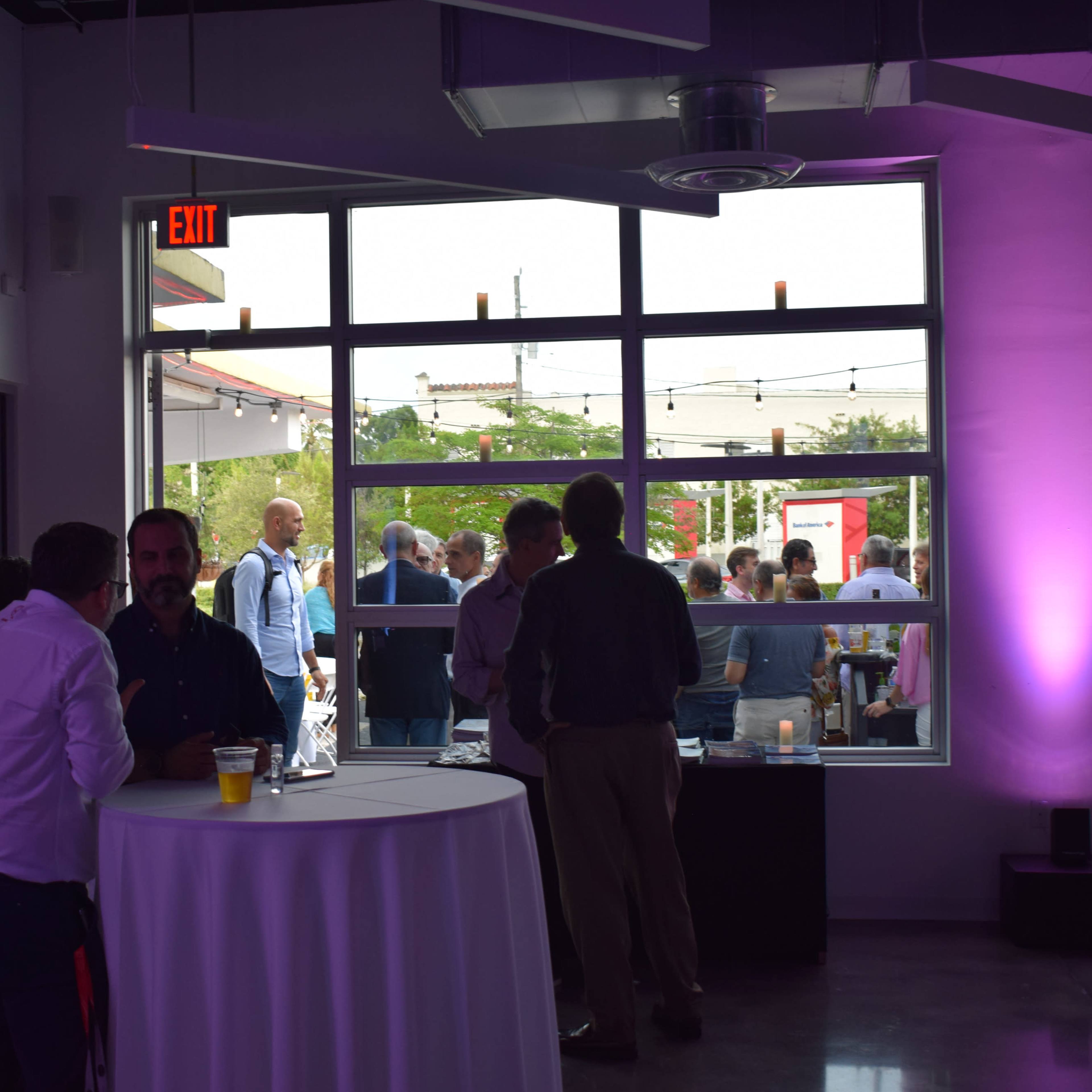 A group of people network in a brightly lit room with large windows overlooking an outdoor gathering.
