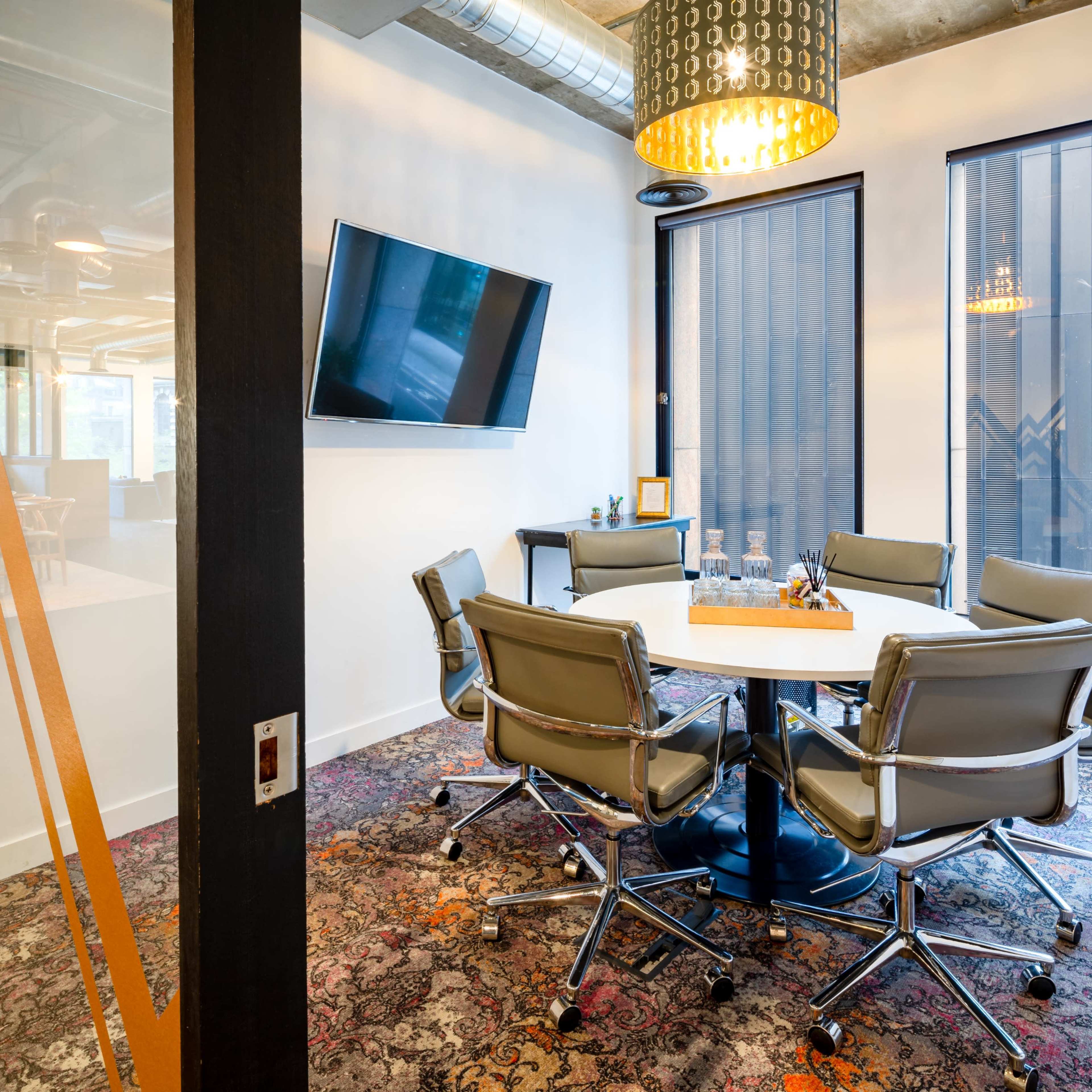 A modern conference room featuring a round table surrounded by ergonomic chairs, a wall-mounted television, and large windows with blinds.