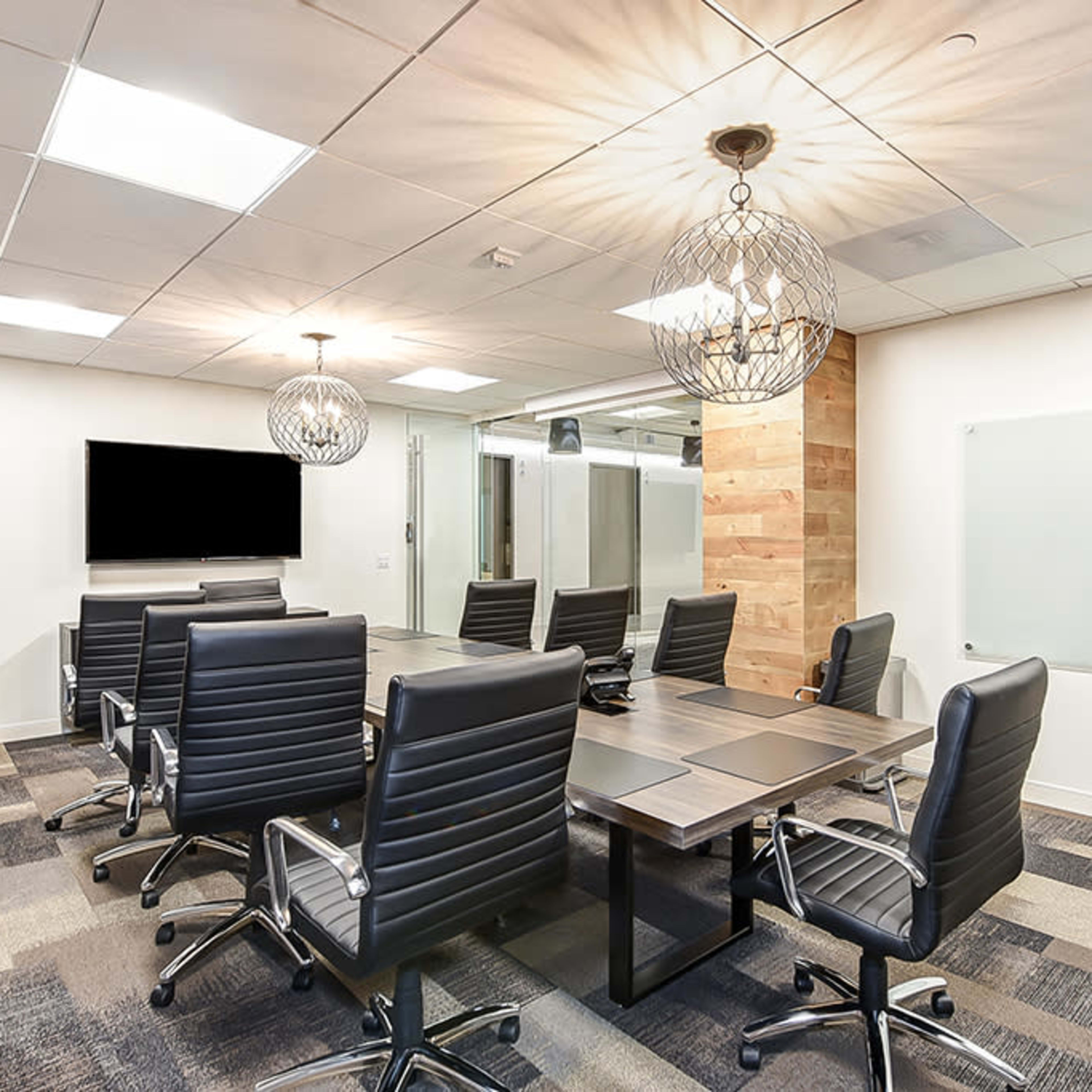 A modern conference room features a long wooden table surrounded by black leather chairs, with pendant lighting and a large screen mounted on the wall.