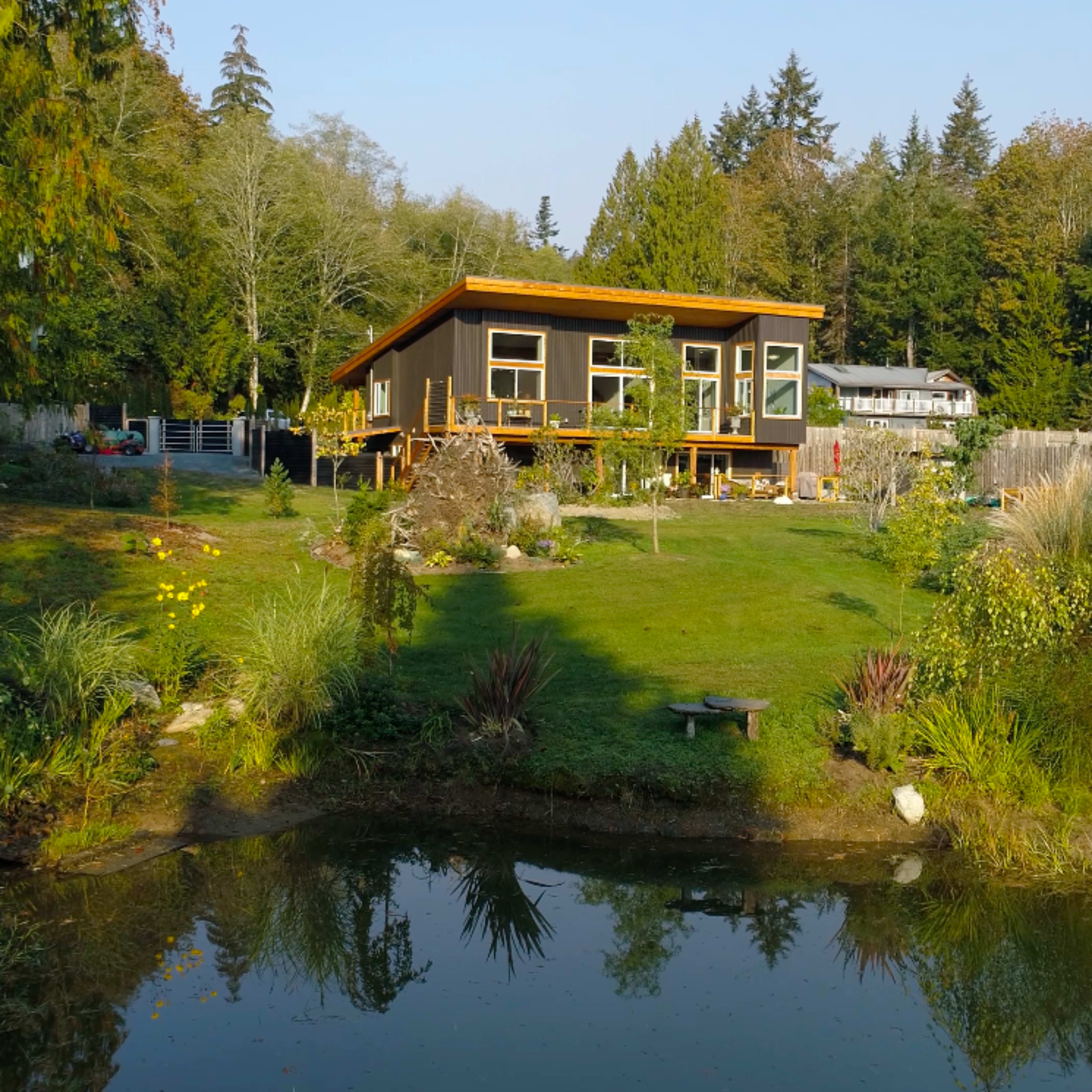 A modern house with large windows is situated on a grassy lawn beside a serene pond, surrounded by trees and landscaping.