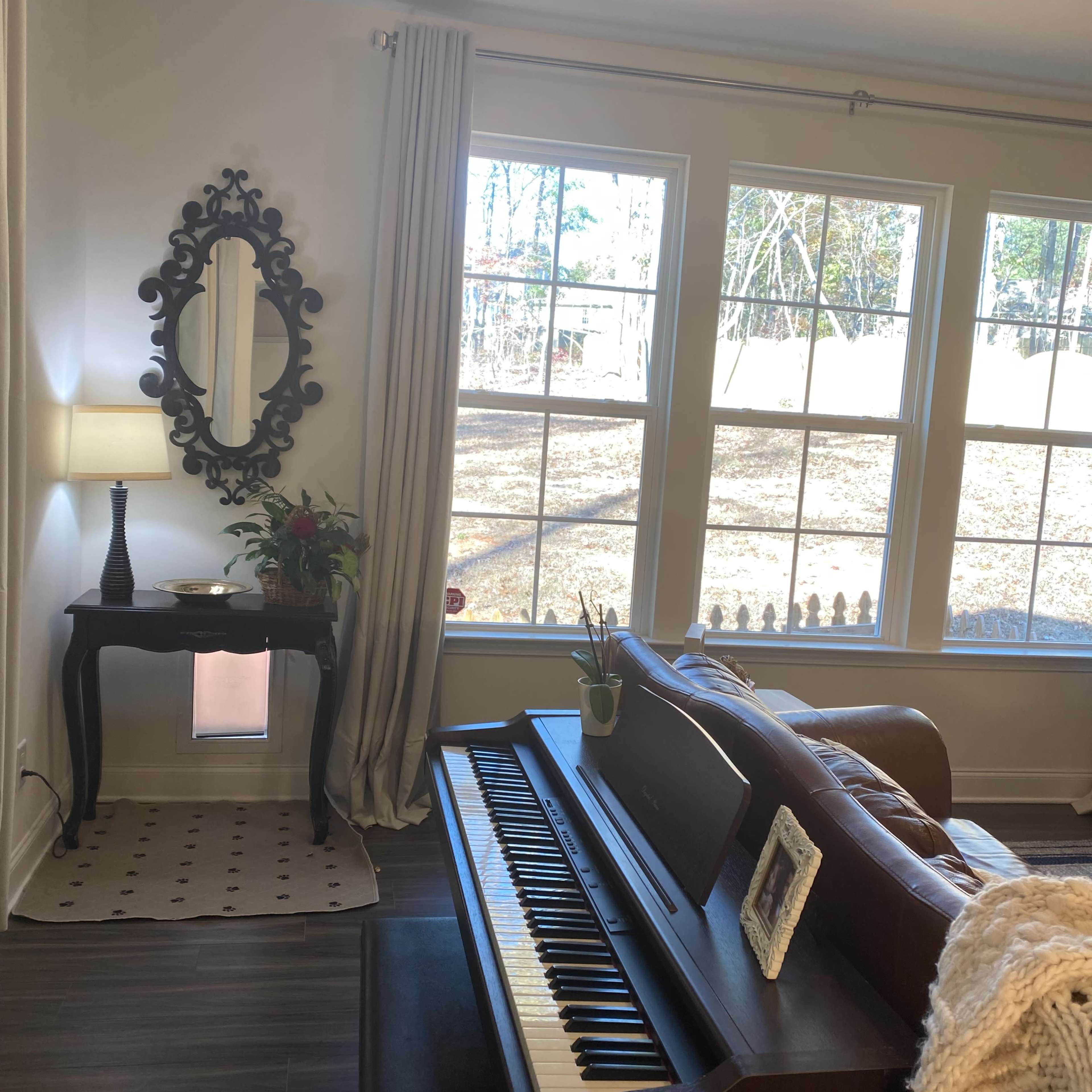 A piano is positioned in a living room with large windows overlooking a backyard, accompanied by a lamp, a mirror, and a small table.