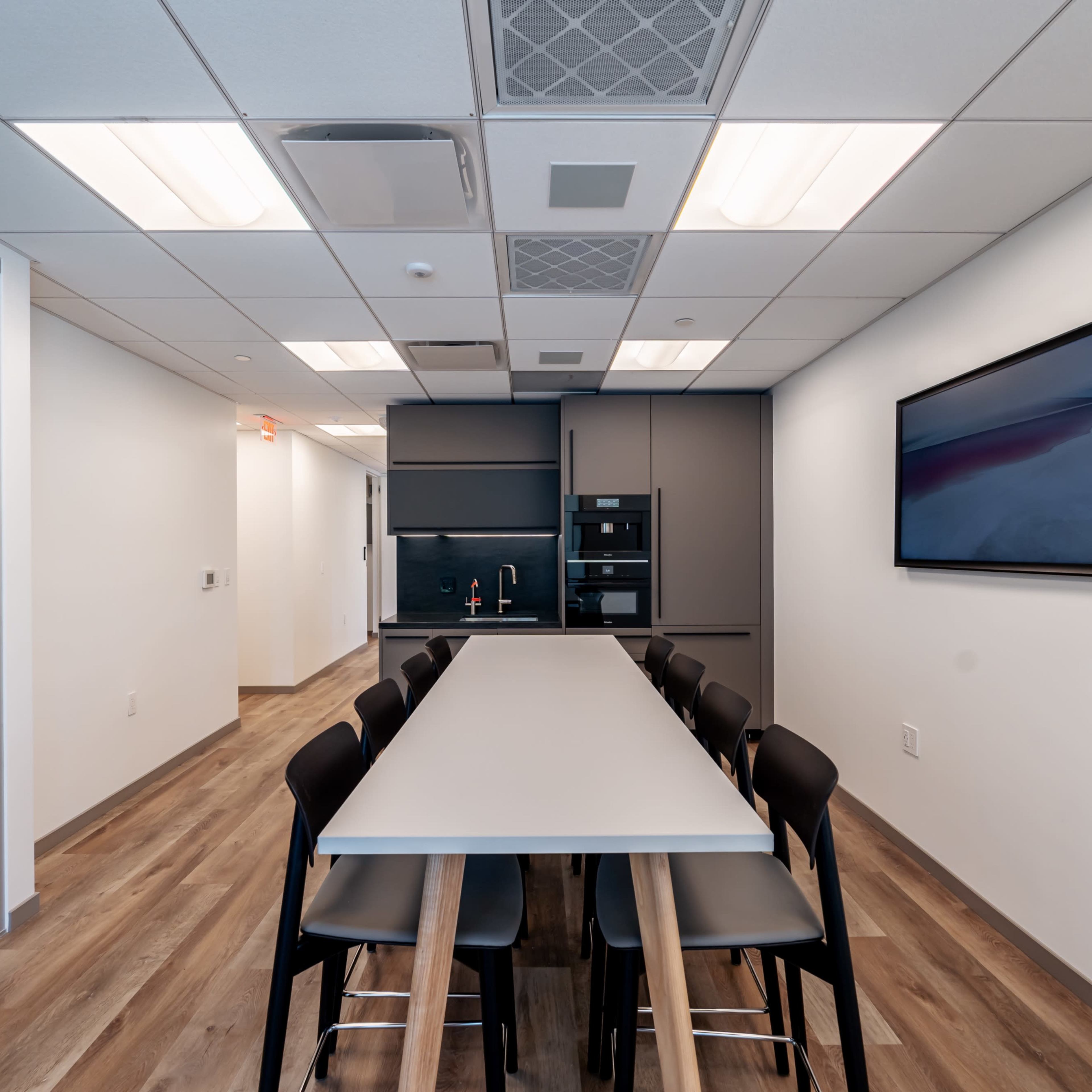 A modern conference room with a long table surrounded by black chairs, a kitchenette in the back, and a wall-mounted screen.