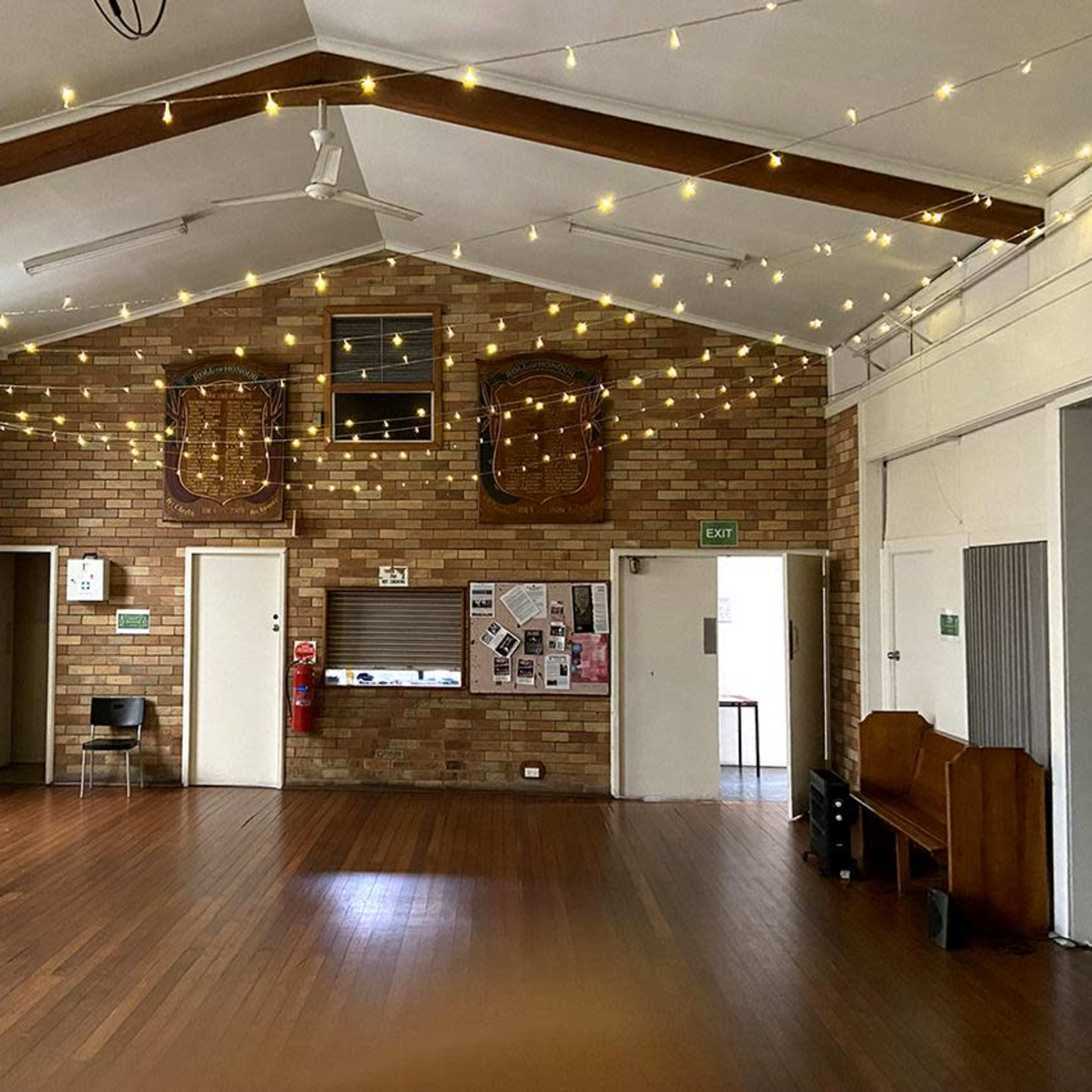 A spacious interior hall with wooden flooring, brick walls, and decorative string lights suspended from the ceiling.