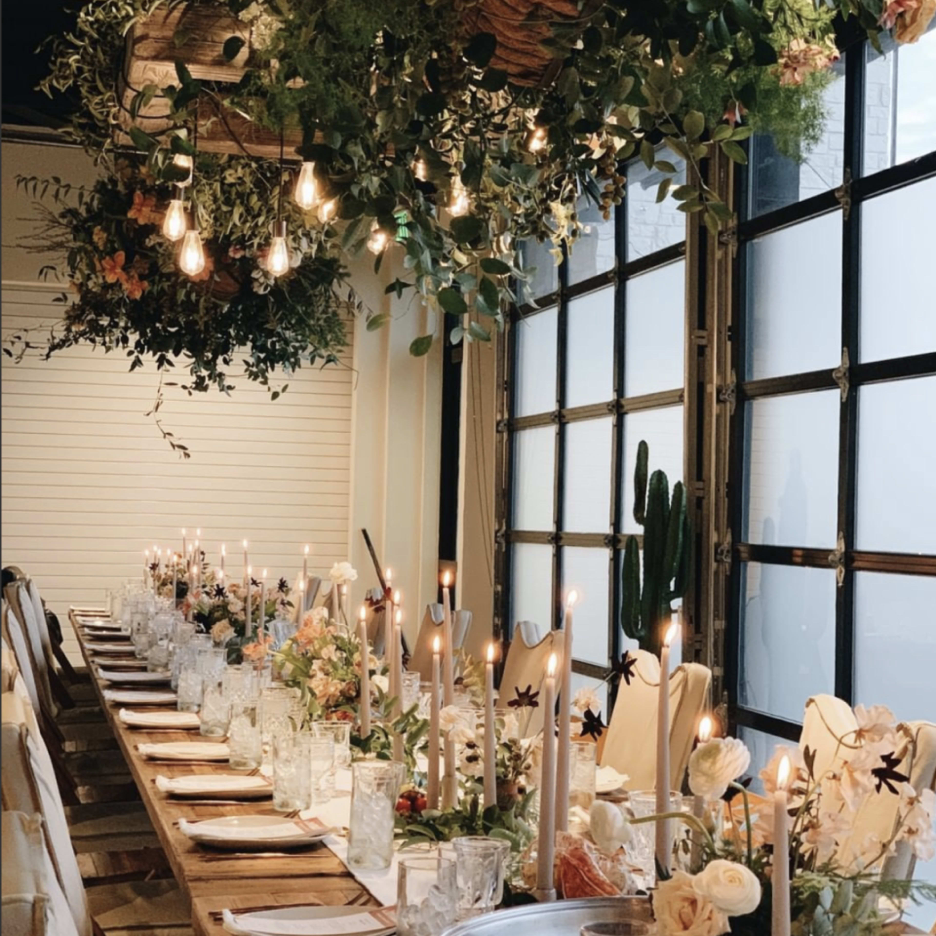 A long dining table, set with candles and floral arrangements, is decorated with hanging plants and pendant lights in a modern venue.