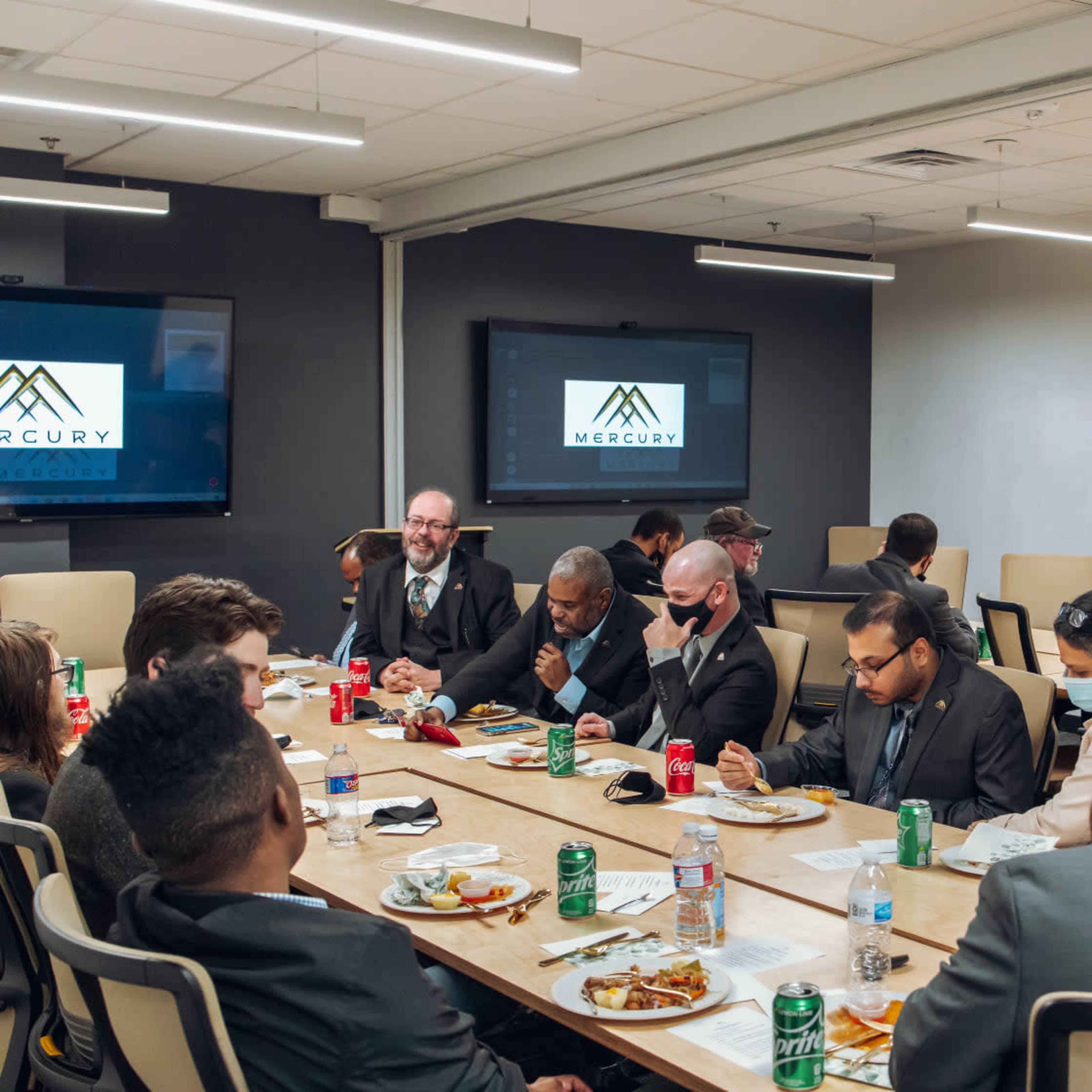 A group of people in business attire are seated around a long table, eating and engaged in conversation during a meal in a conference room.