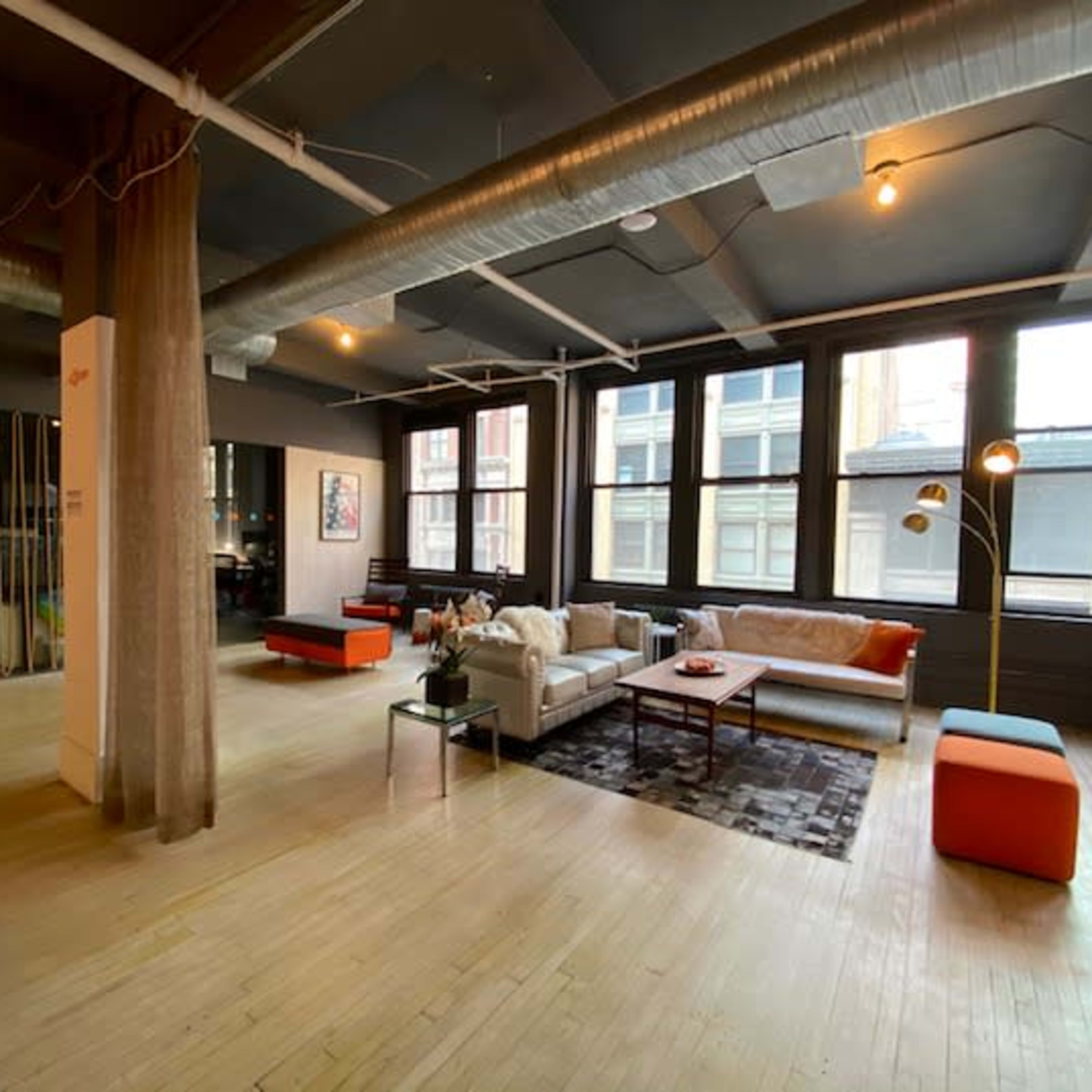 A spacious living room with large windows, modern furniture including a sofa, a coffee table, and accent chairs, along with hardwood flooring and exposed ductwork.