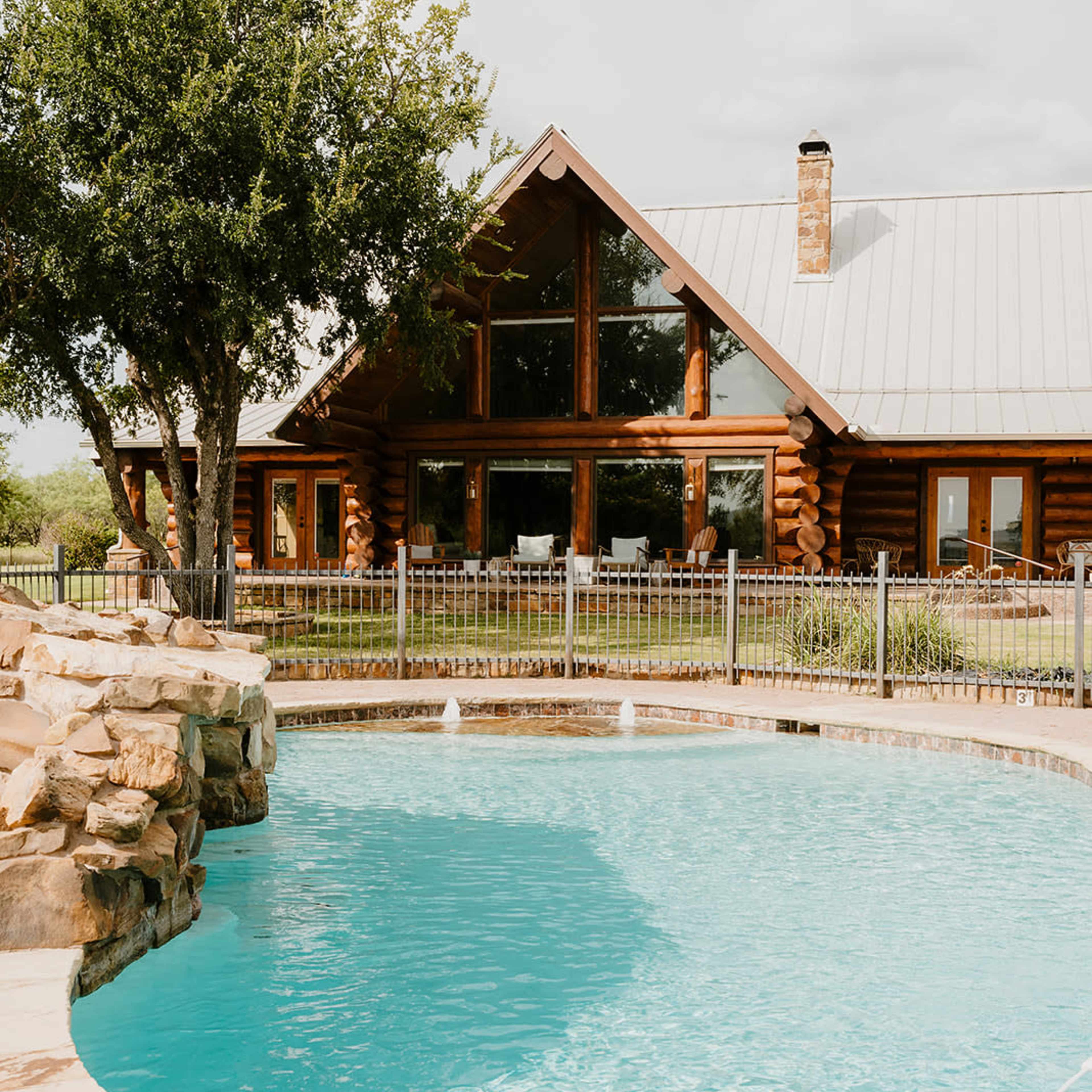 A log cabin with large windows overlooks a blue swimming pool surrounded by a stone patio and lush greenery.