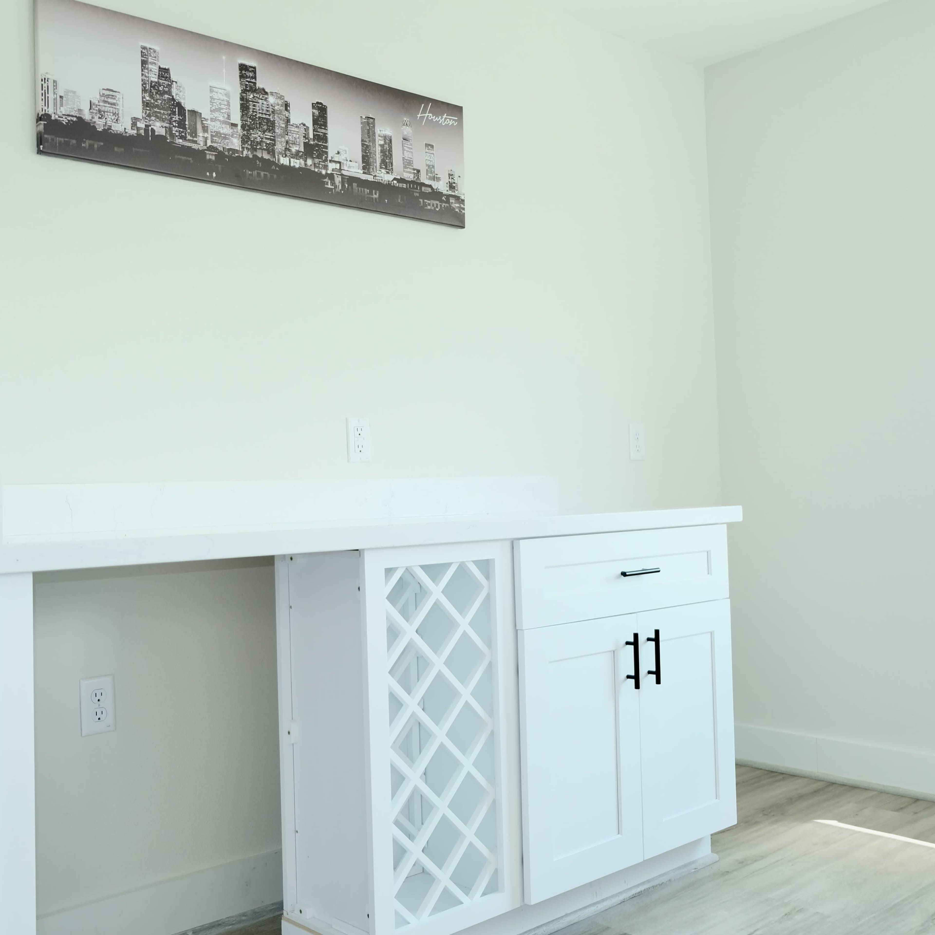 A minimalist workspace featuring a white cabinet with a wine rack and a sleek countertop, accompanied by a black and white cityscape artwork on the wall.