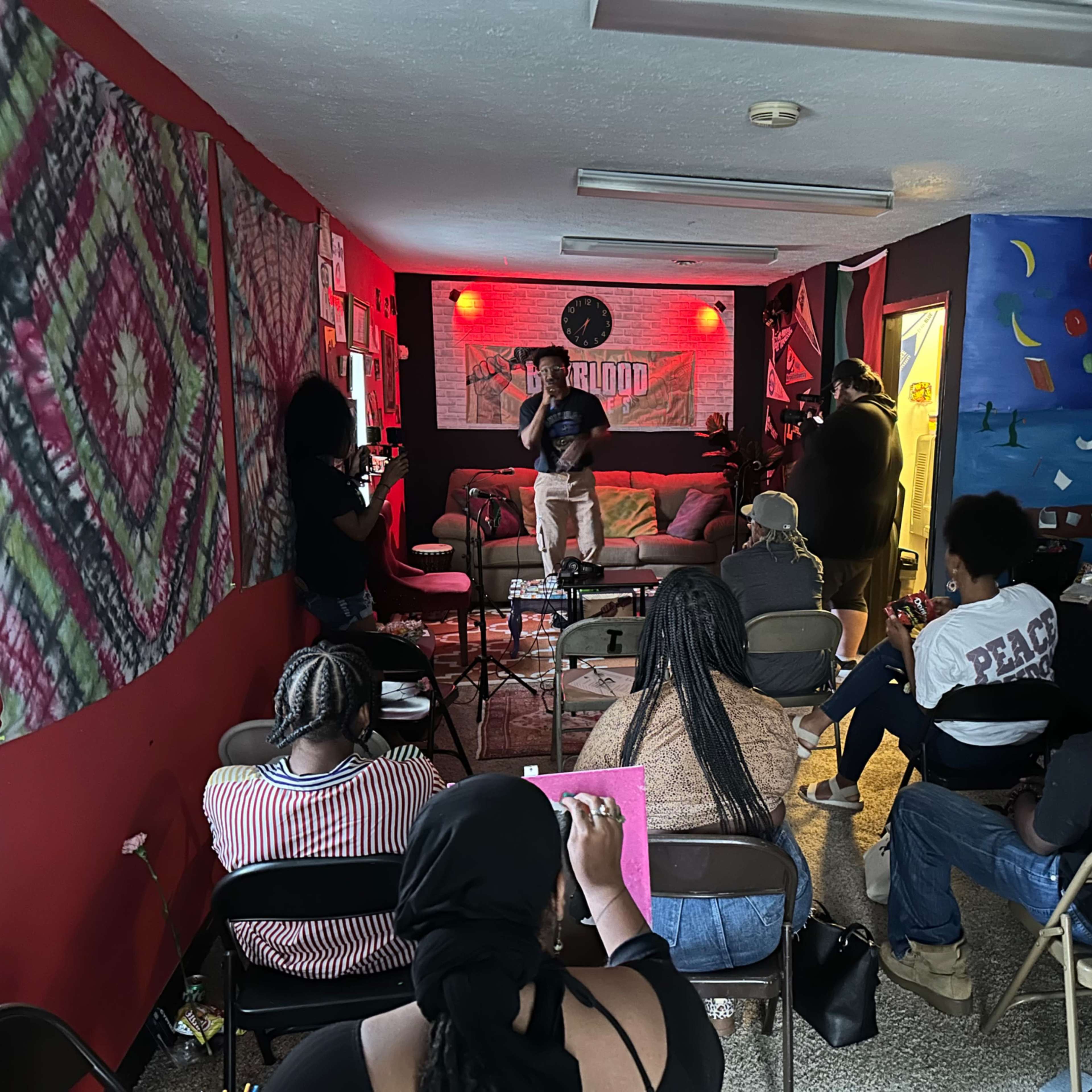A group of people sits in chairs facing a small stage in a colorful room decorated with various artworks.