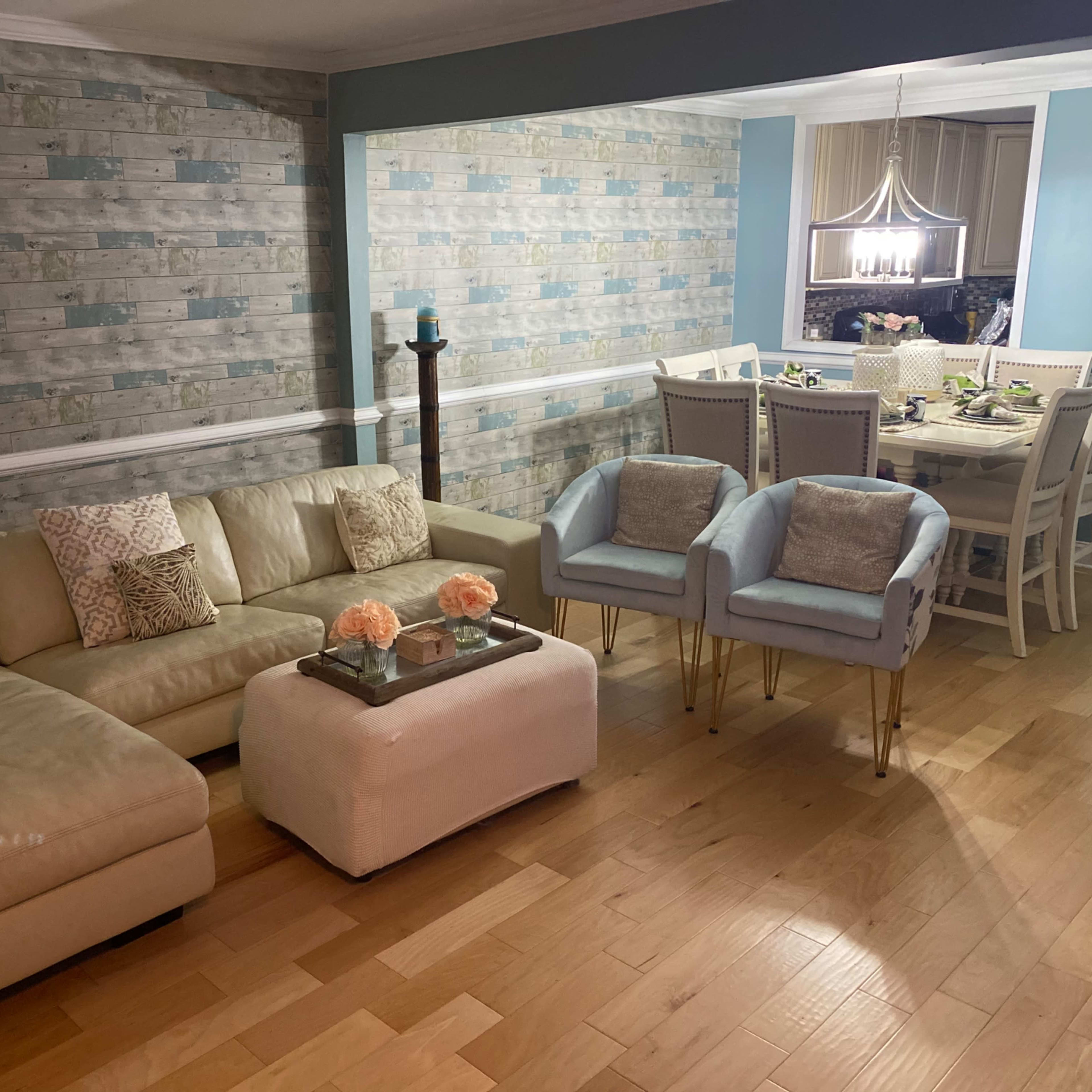 A modern living room featuring a beige sectional sofa, a small white ottoman with decorative flowers, and two light blue armchairs, with a dining area visible in the background.