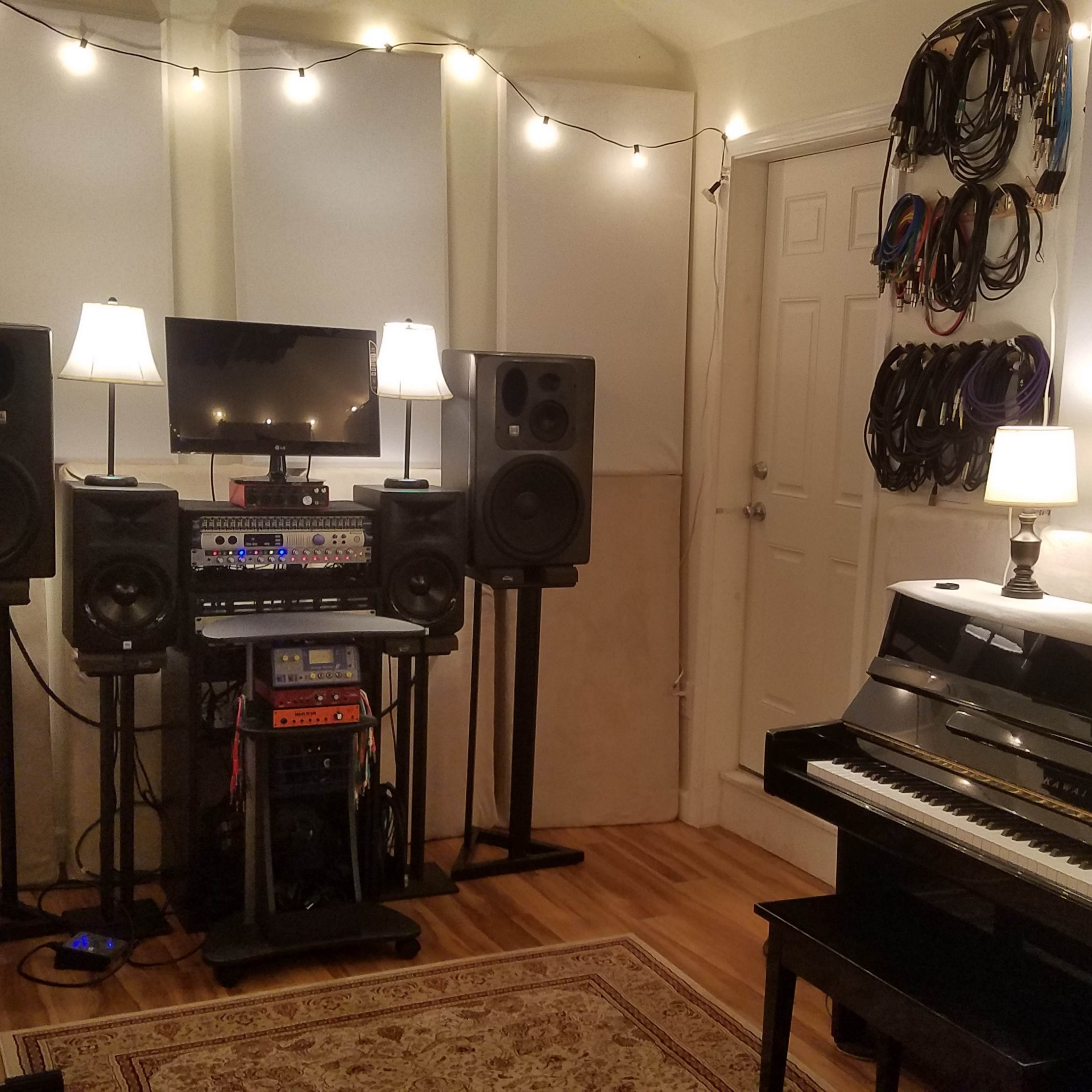 A music studio with a digital piano, an acoustic piano, audio speakers, a mixing console, and ambient lighting.
