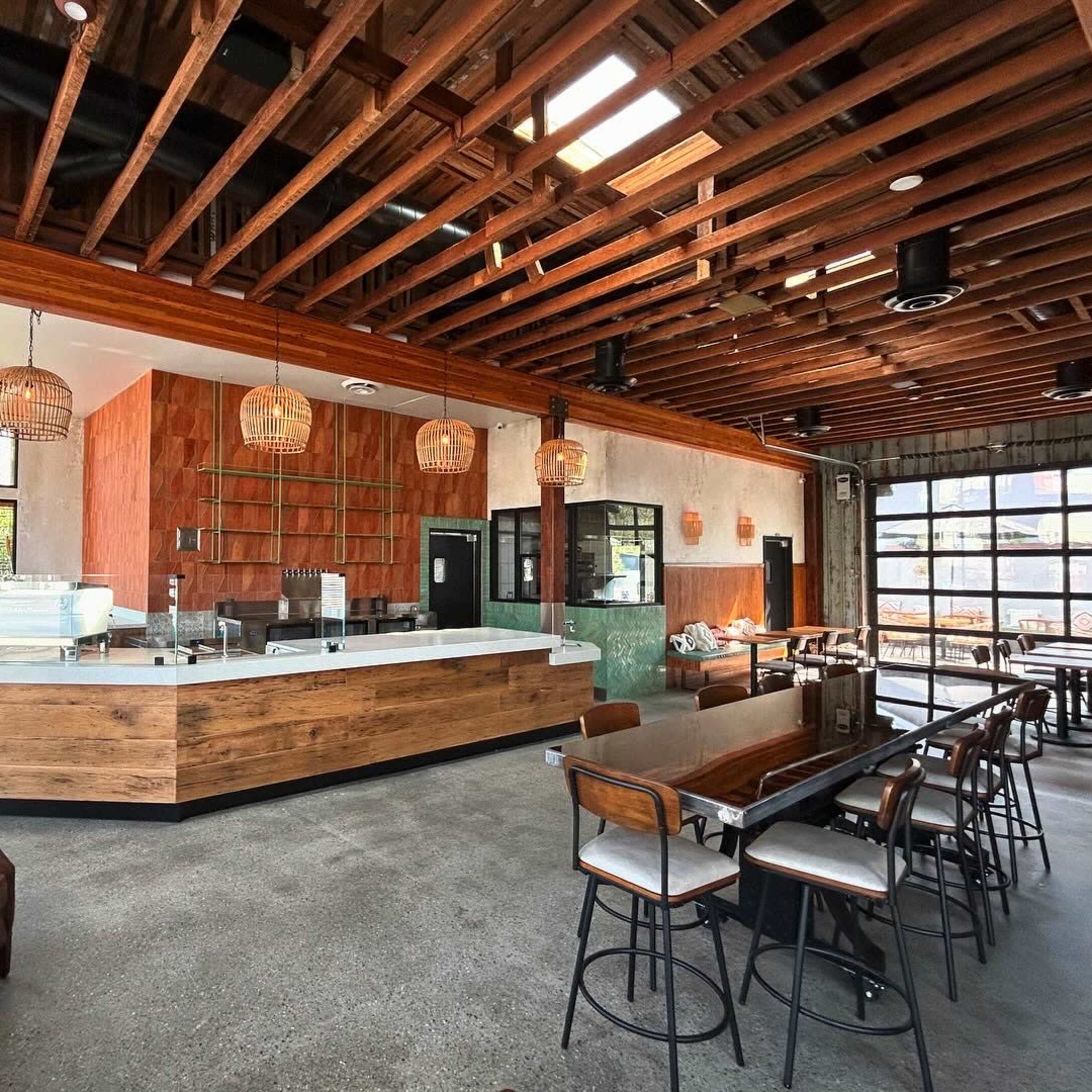 A modern café interior featuring a wooden counter, bar seating, and overhead wooden beams with large windows allowing natural light.