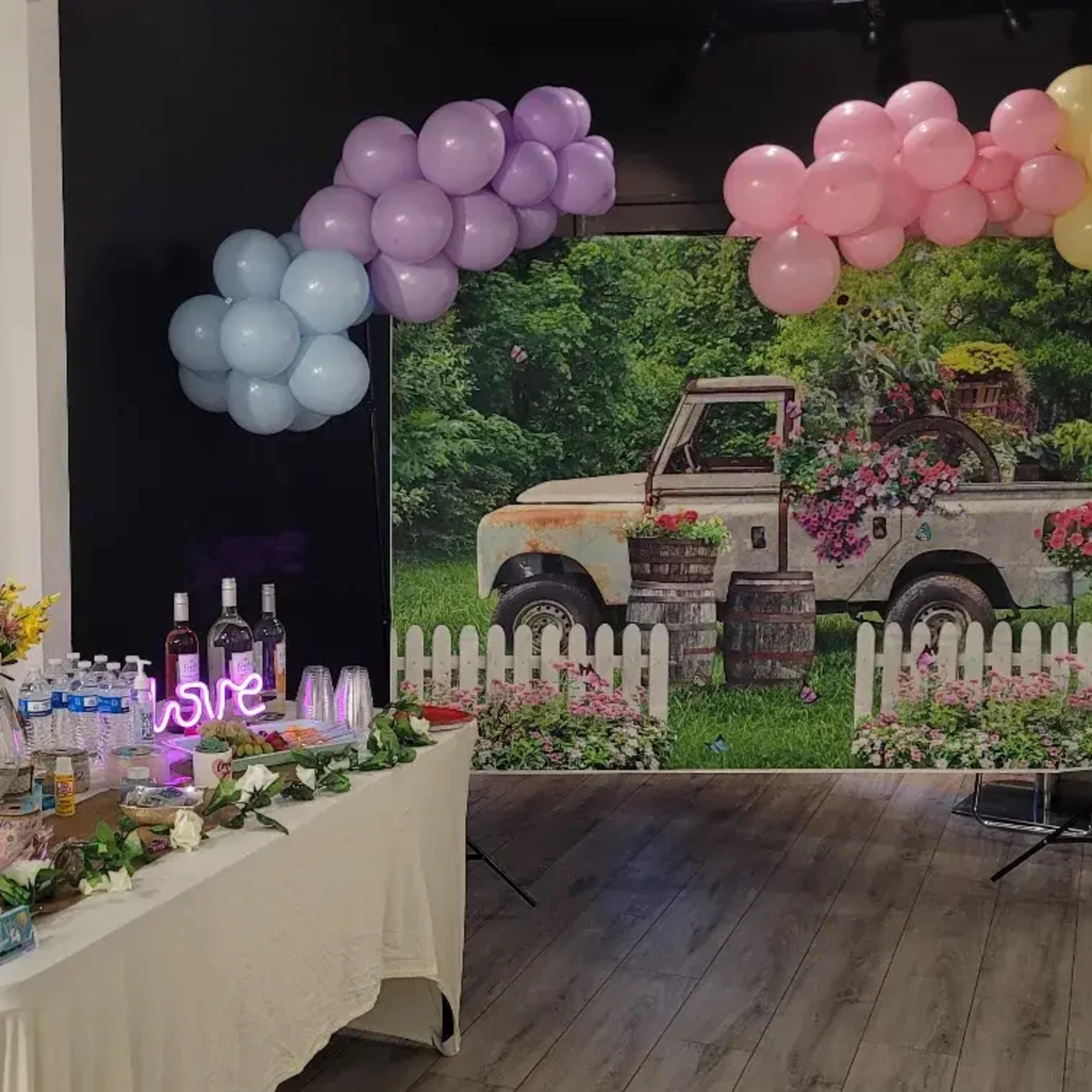 A decorated table with drinks and flowers is positioned in front of a backdrop featuring a vintage truck surrounded by greenery and balloons of various colors.