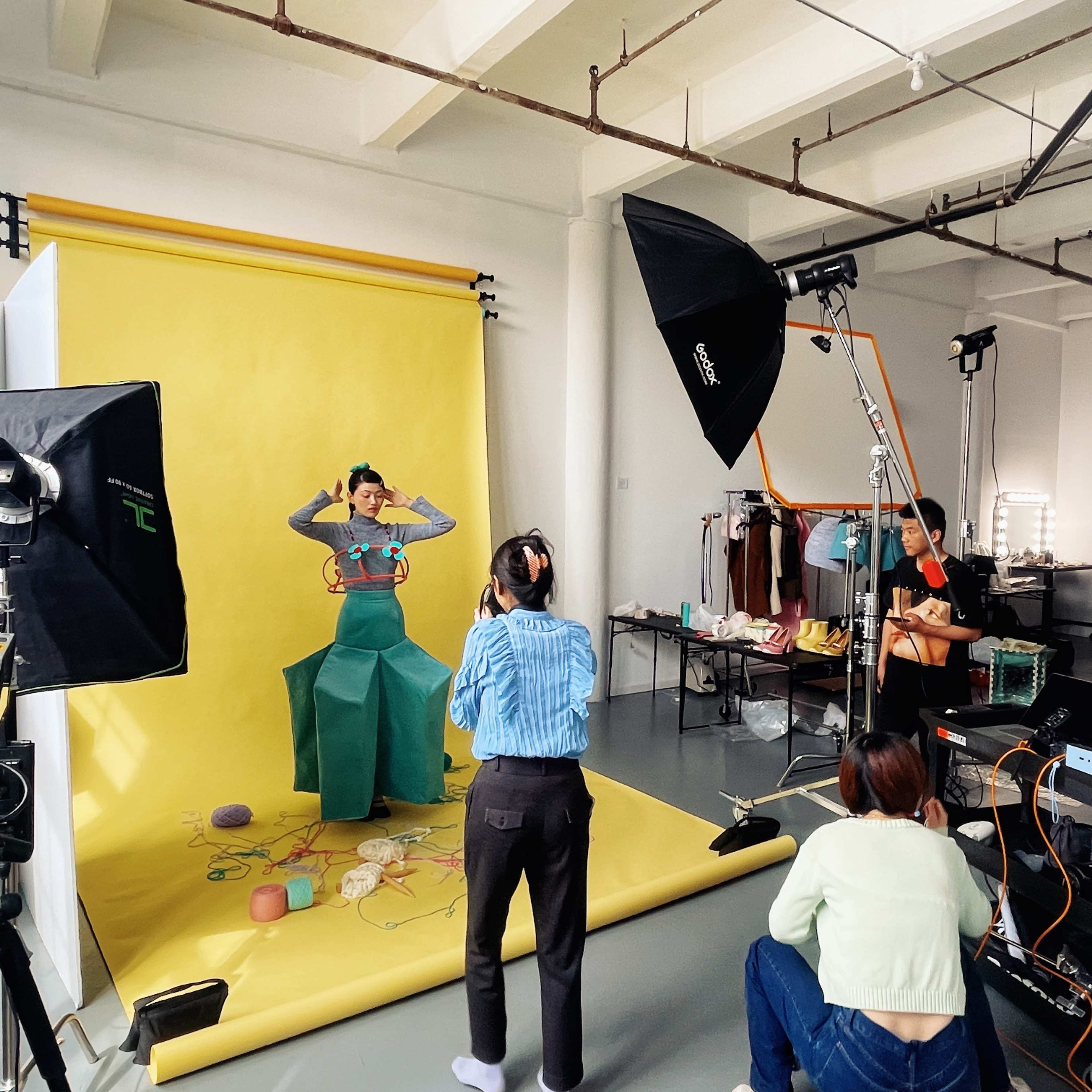 A model poses in front of a yellow backdrop while a team of photographers and assistants work in a studio setting.