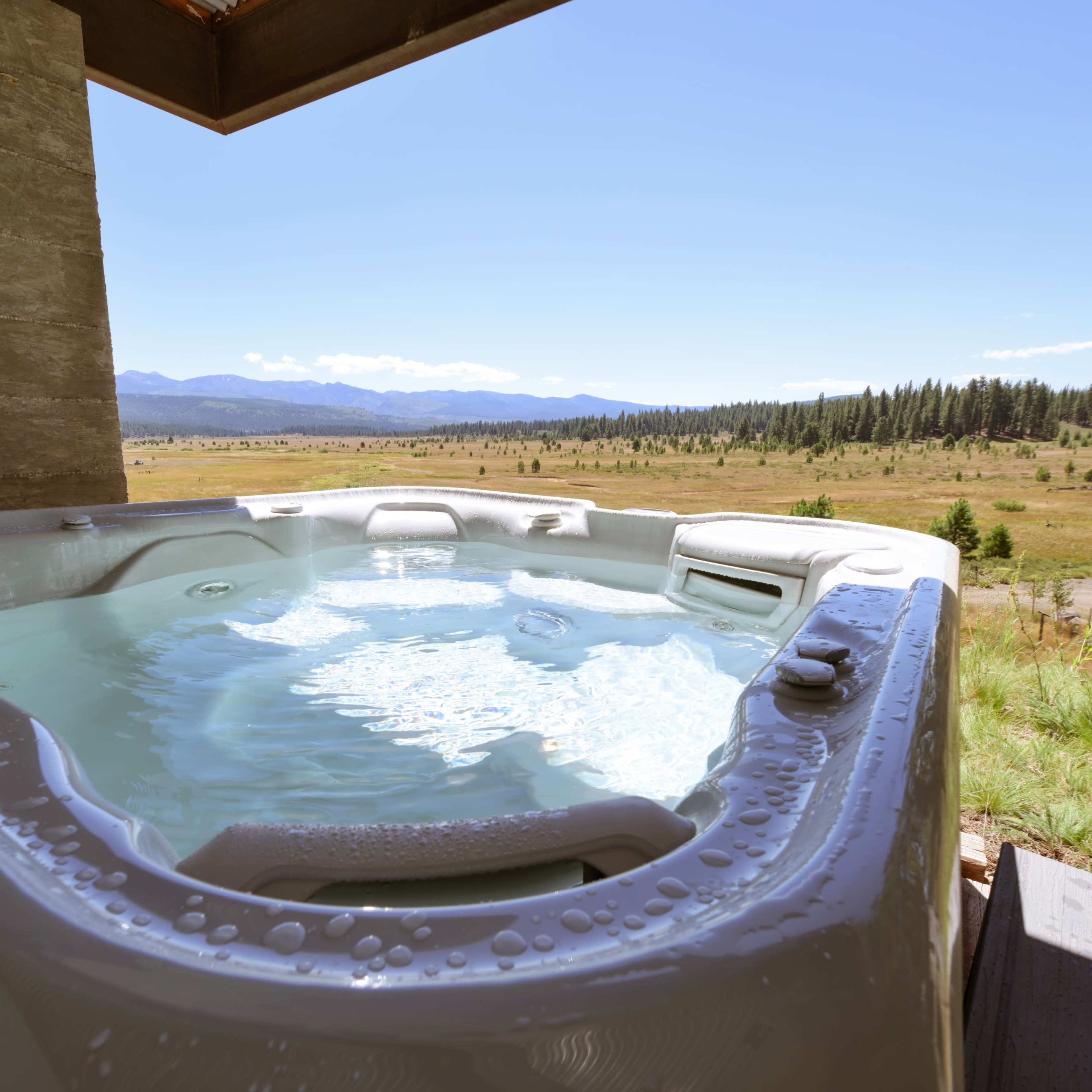A hot tub filled with water sits on a deck overlooking a scenic landscape of mountains and fields under a clear blue sky.