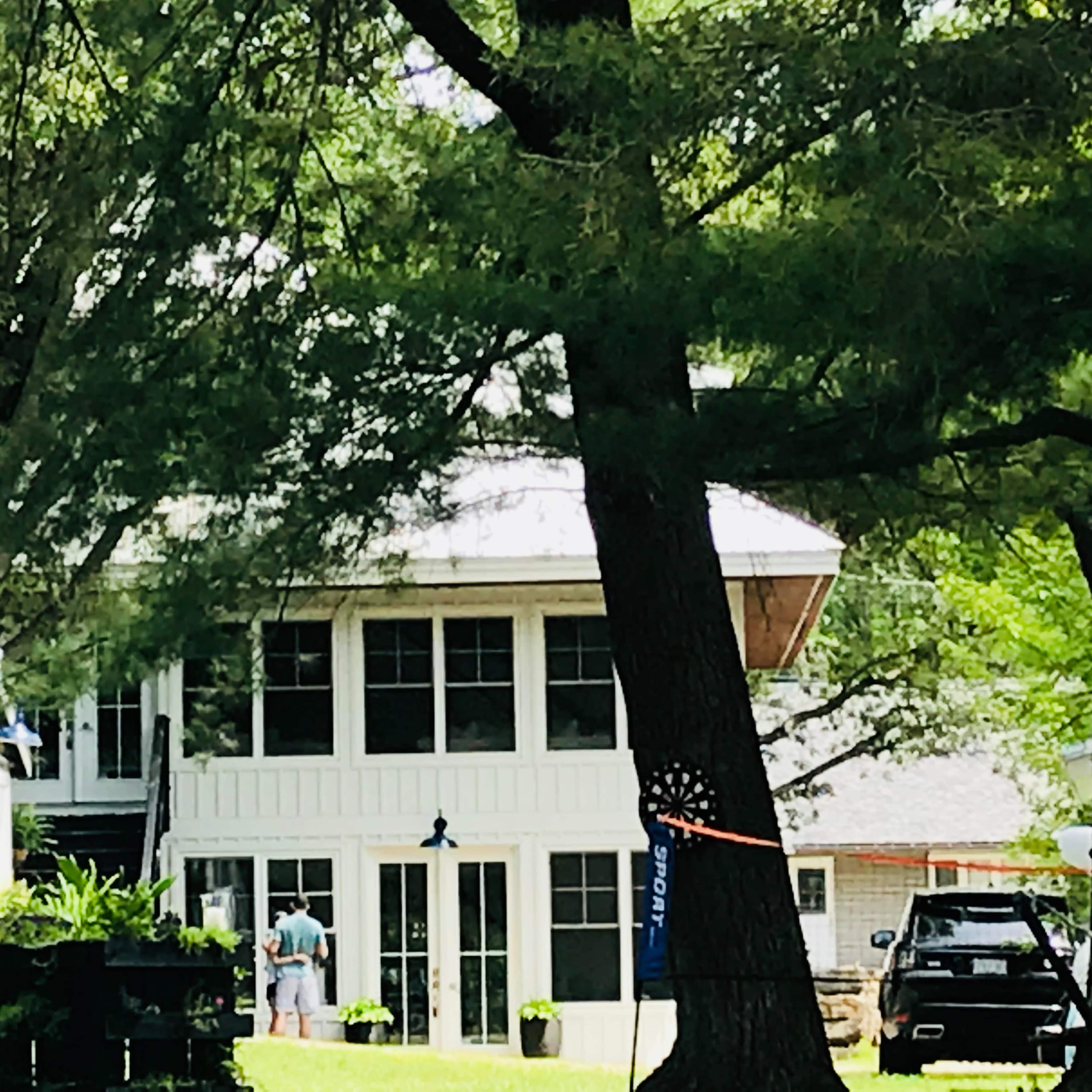 A two-story house with a white exterior and large windows is partially obscured by trees, while two people stand in front of it.