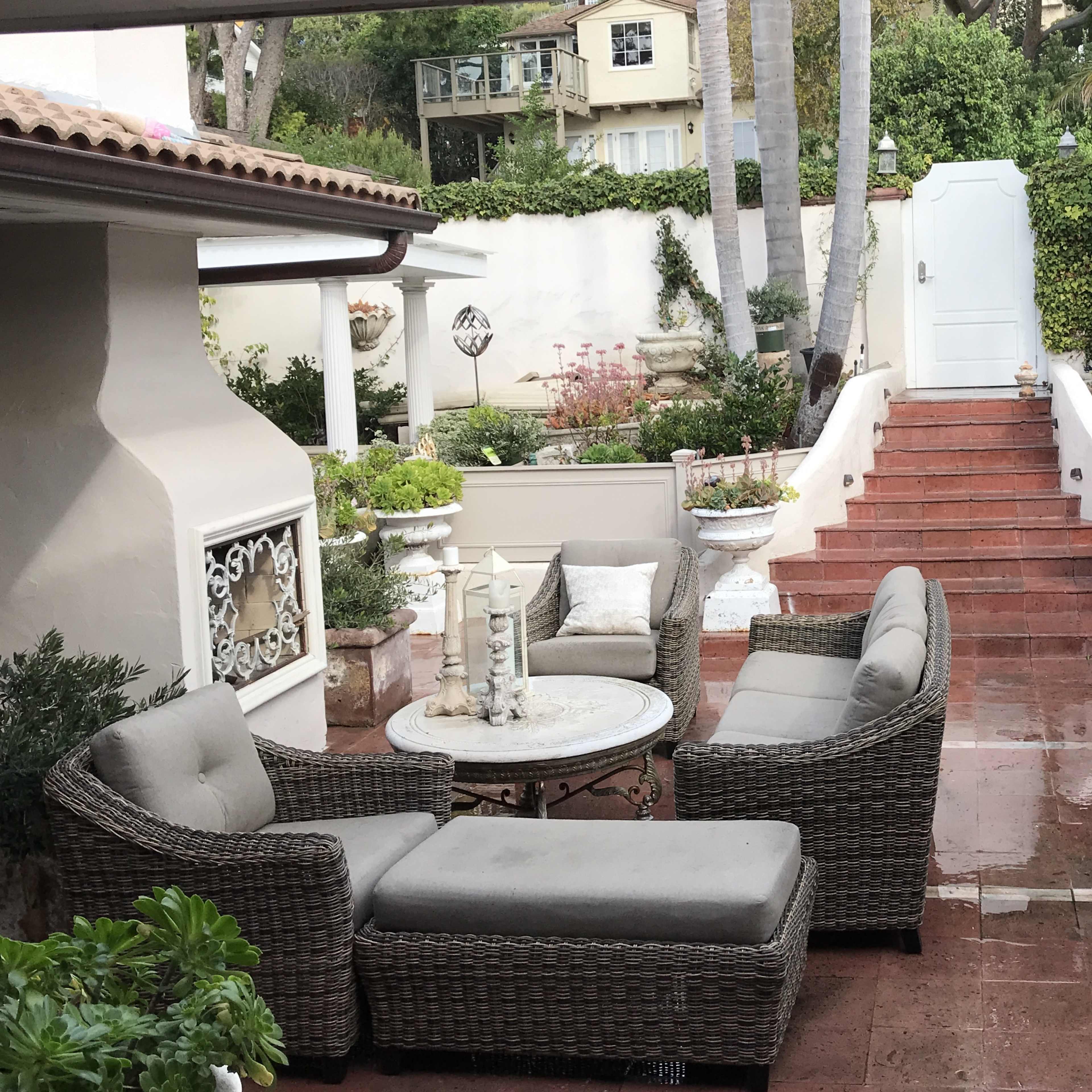 A patio area features wicker furniture and a stone table, with steps leading up to a garden flanked by palm trees and decorative elements.
