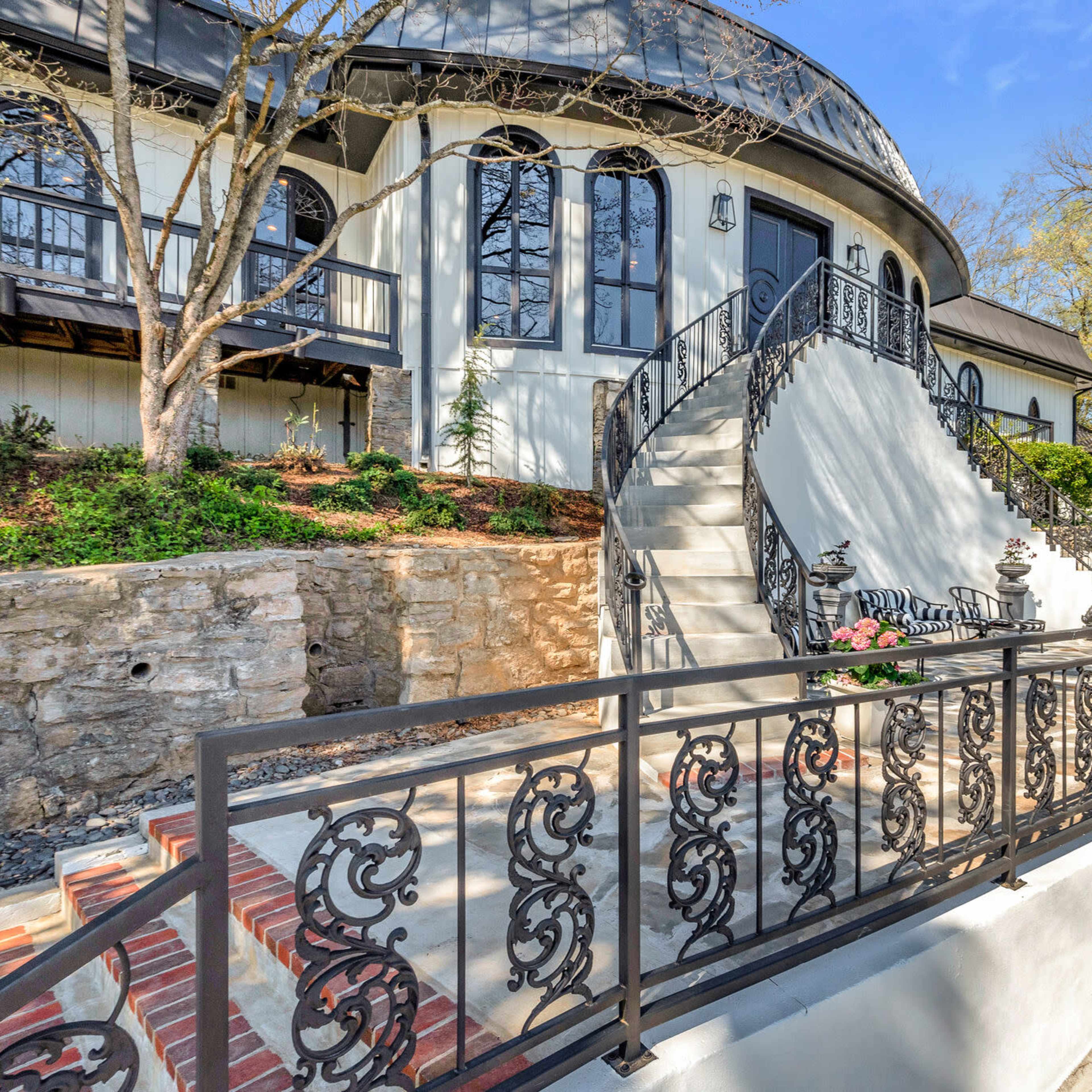 A modern house with a circular design, featuring a stone staircase leading up to an ornate black railing and landscaped surroundings.