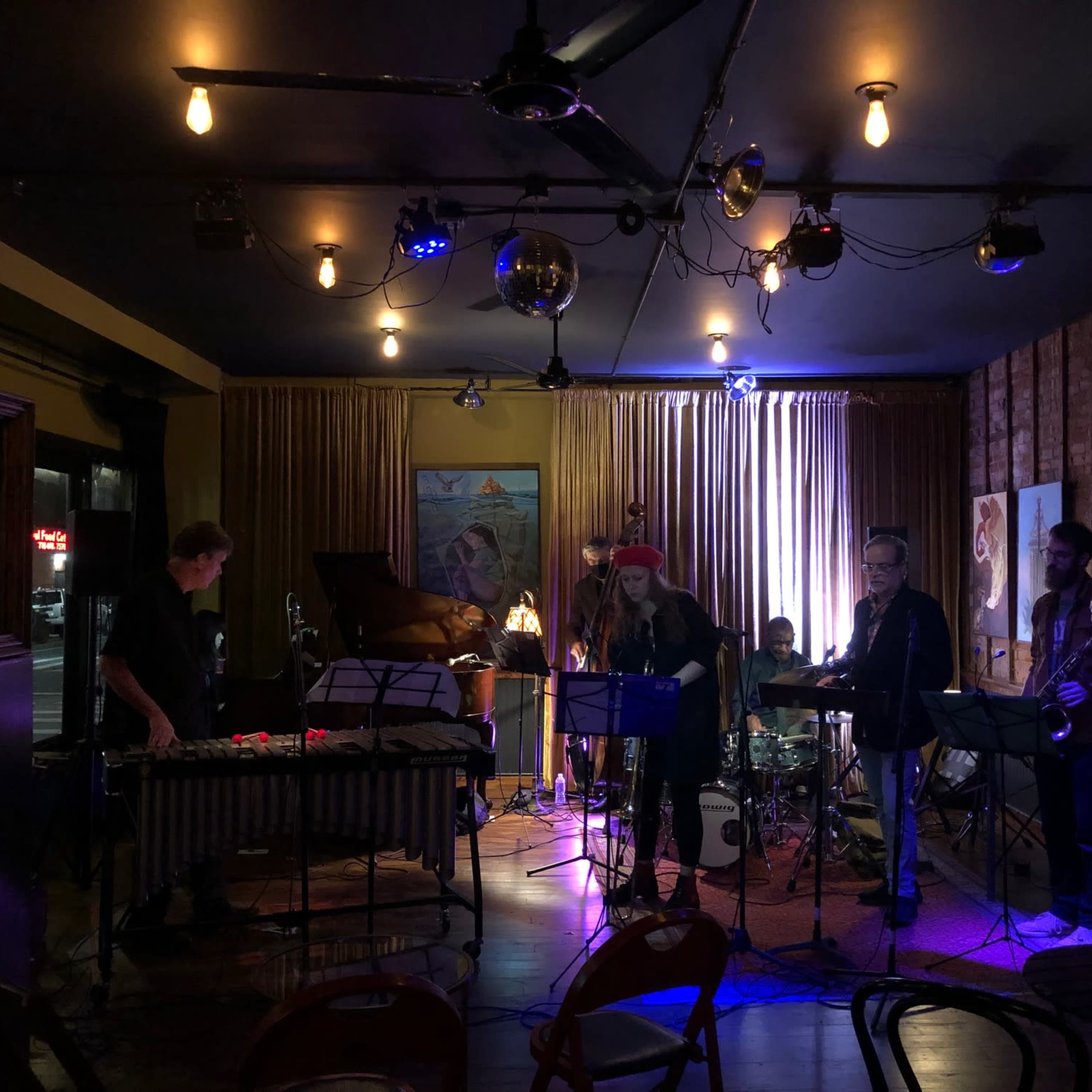 A group of musicians performs on stage in a dimly lit venue, surrounded by instruments and colorful artwork on the walls.