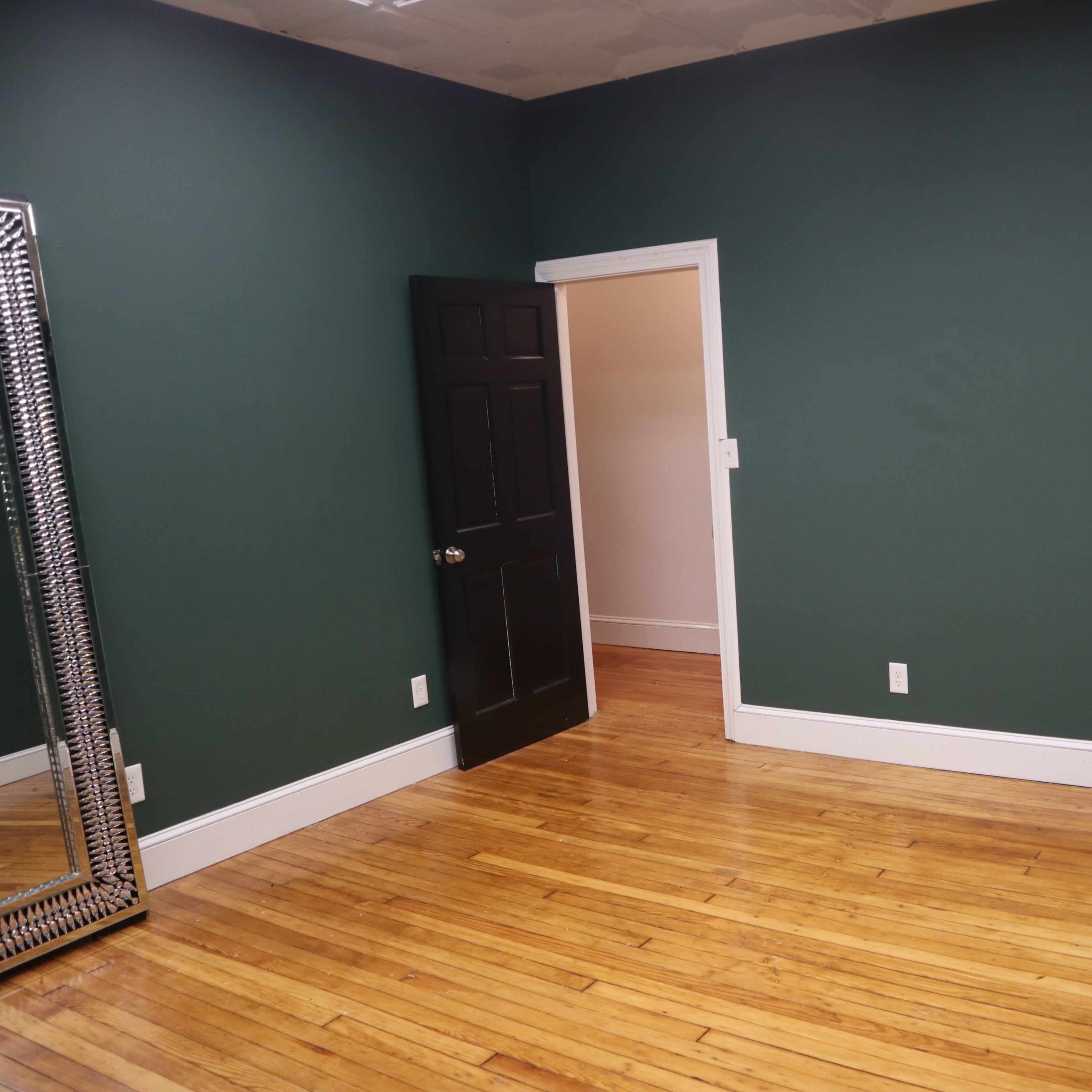 A room with dark green walls, a wooden floor, a large mirror on one side, and a black door leading to another space.