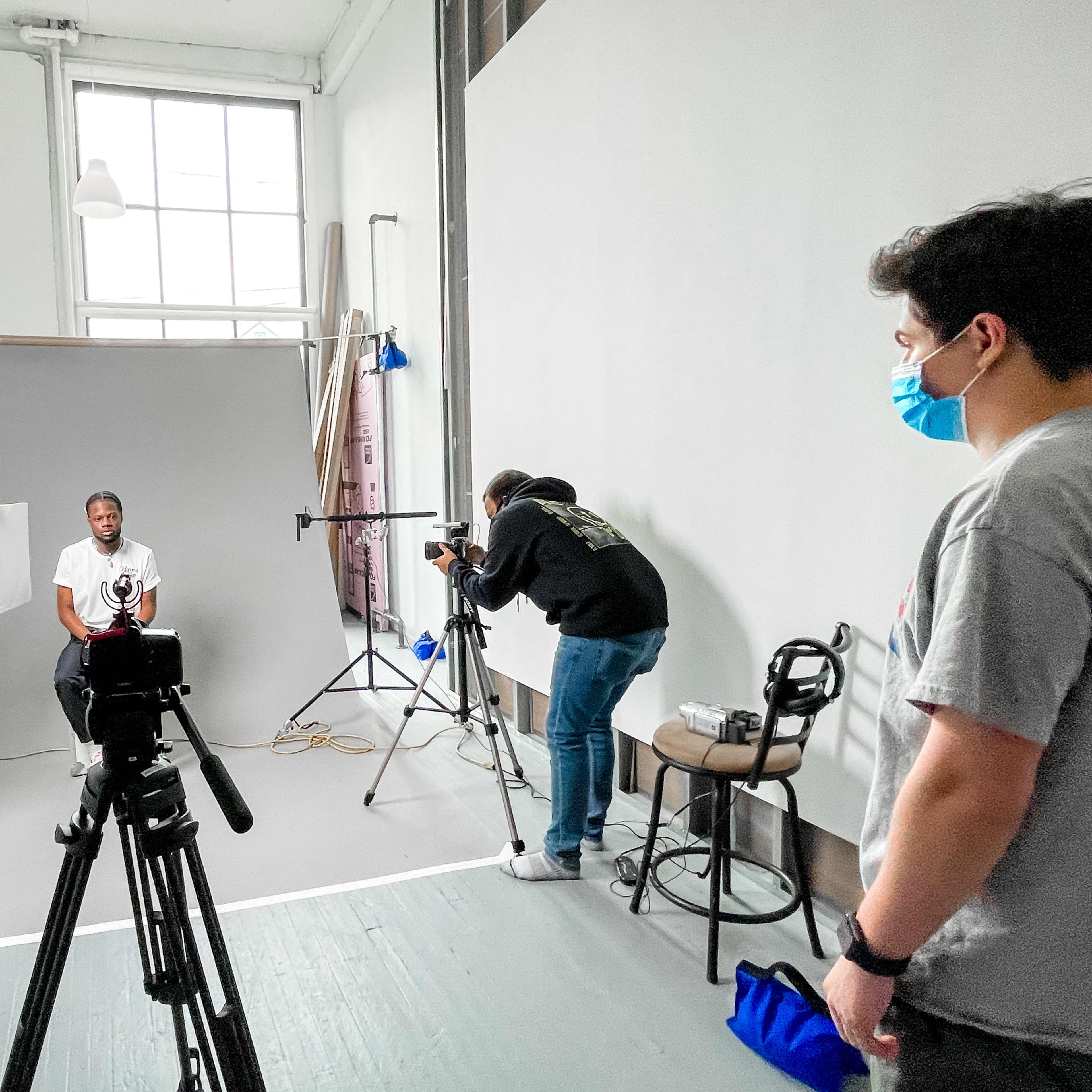 A photoshoot is taking place in a studio, featuring a seated subject and a team of individuals using camera equipment and lighting setup.