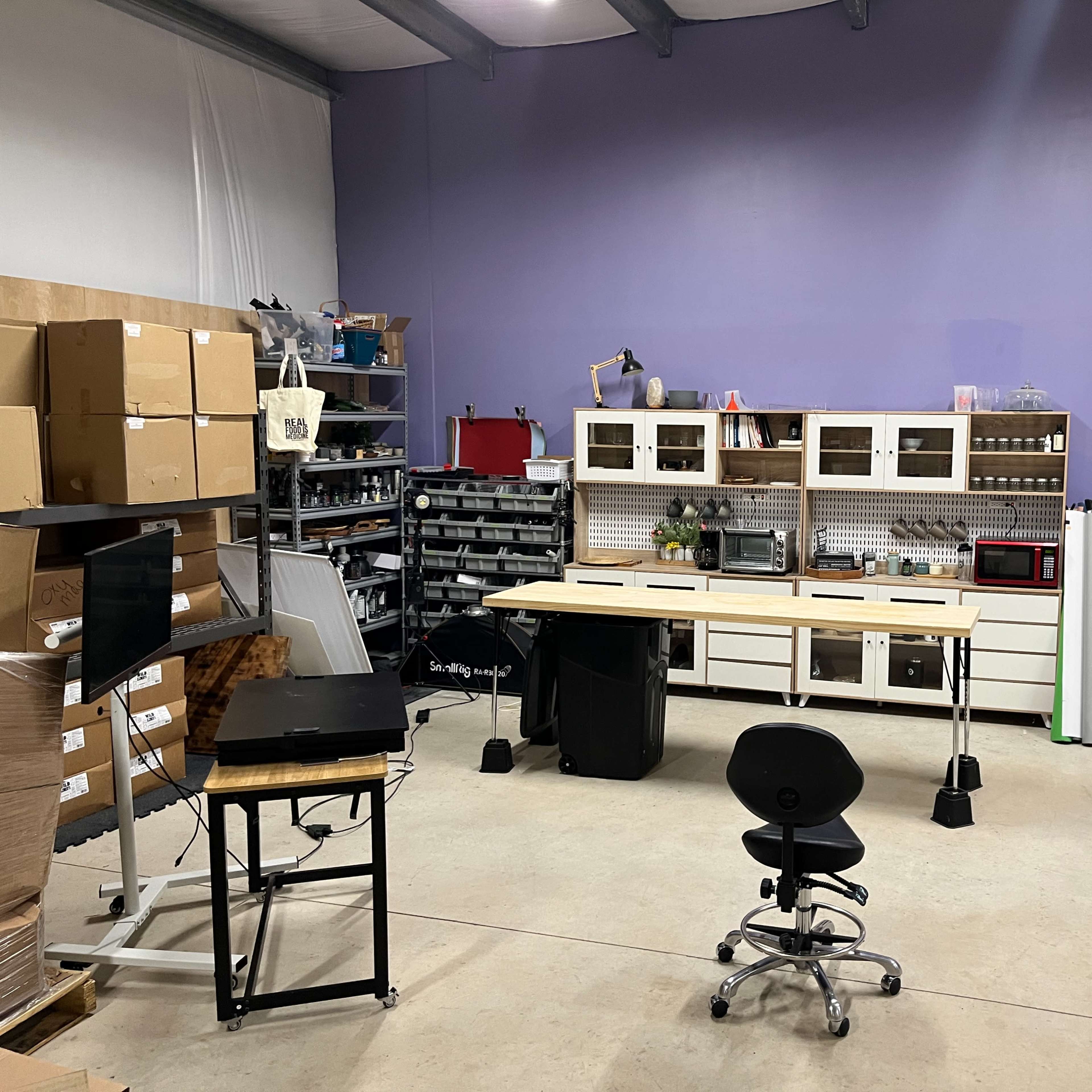 A workspace featuring a central table with stools, storage cabinets, and various tools, set against a purple wall.