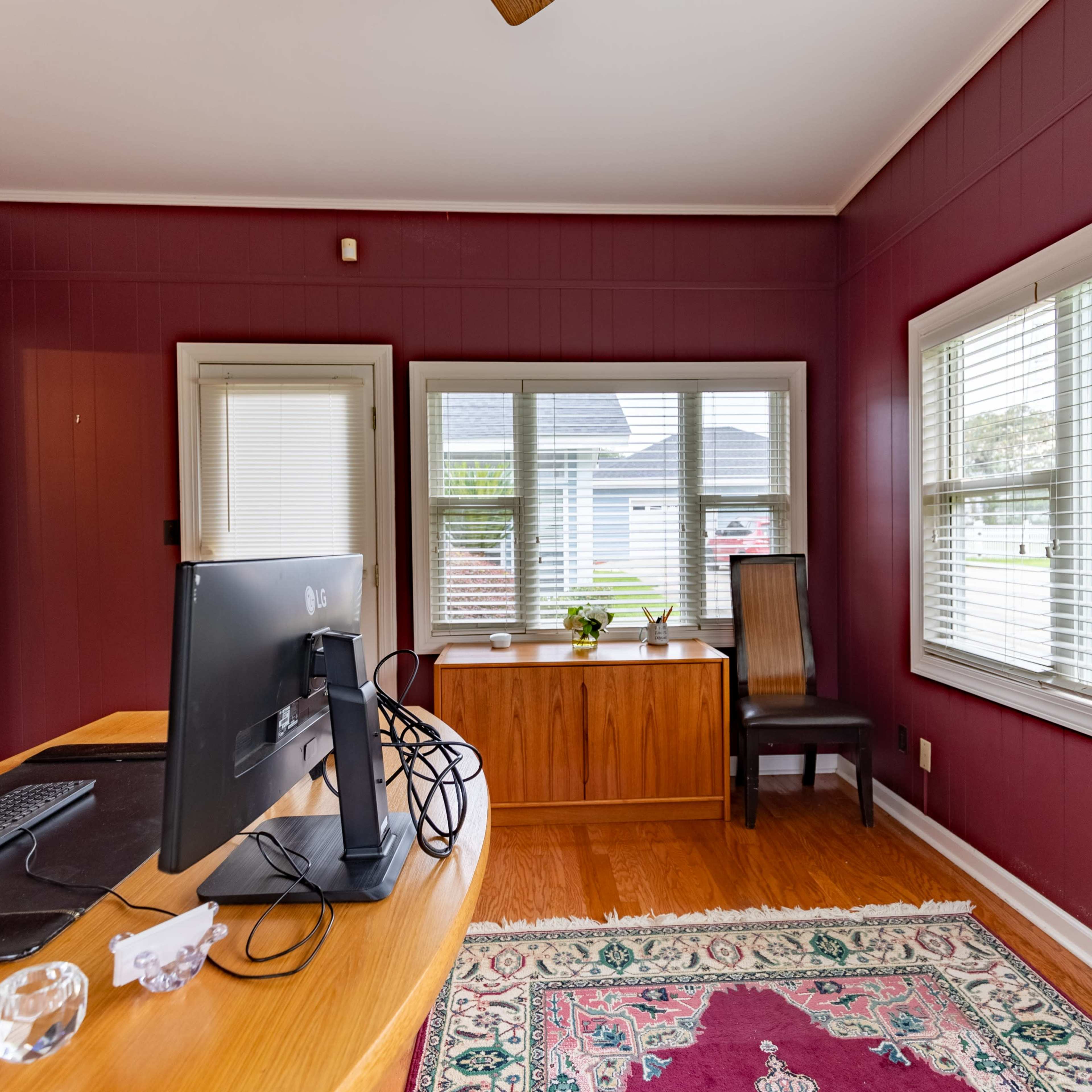 A small office space with a wooden desk, a black computer monitor, and large windows facing an outdoor view.