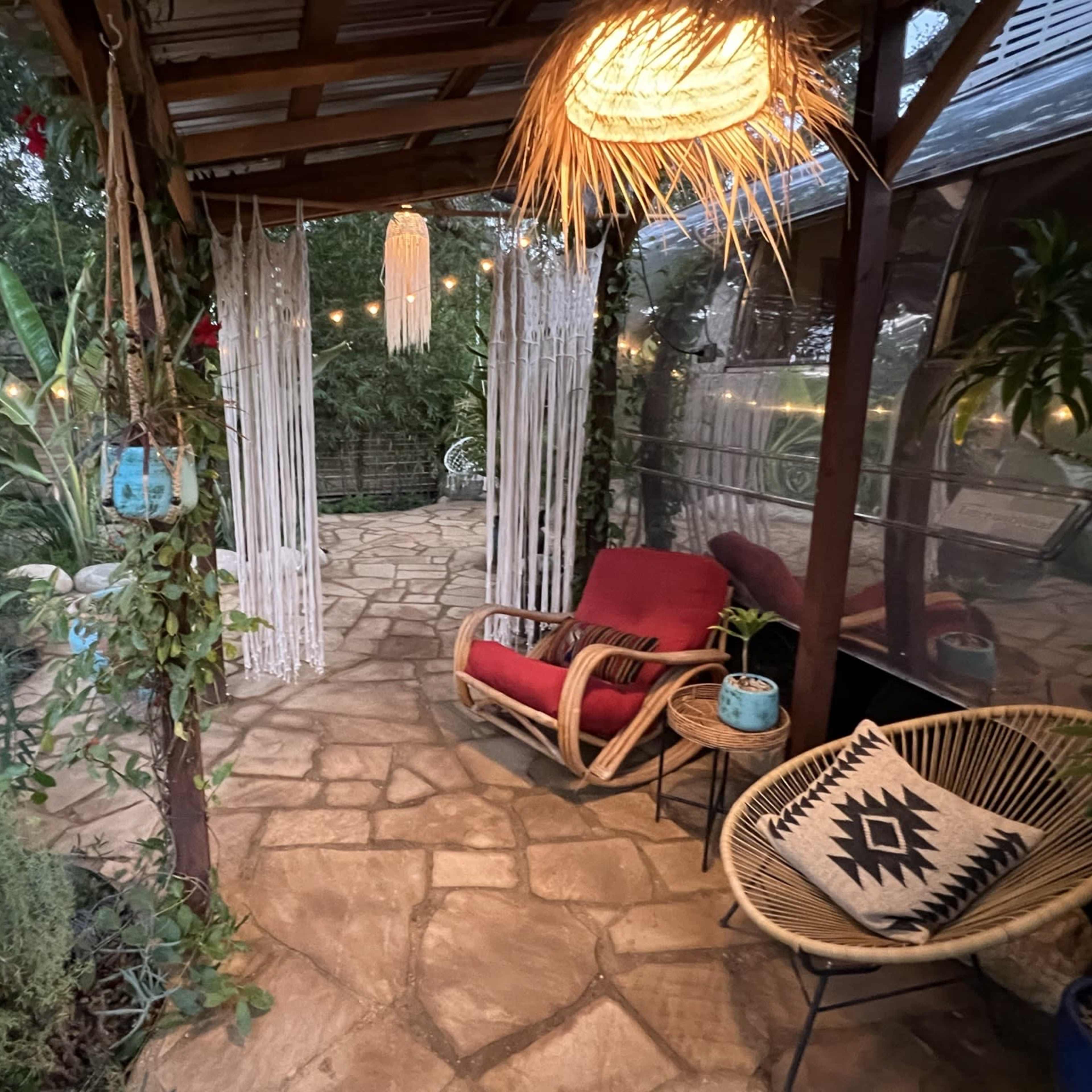 A cozy outdoor patio area with a stone path, surrounded by plants, lit by string lights and lanterns, and furnished with a red chair and a patterned cushion chair.