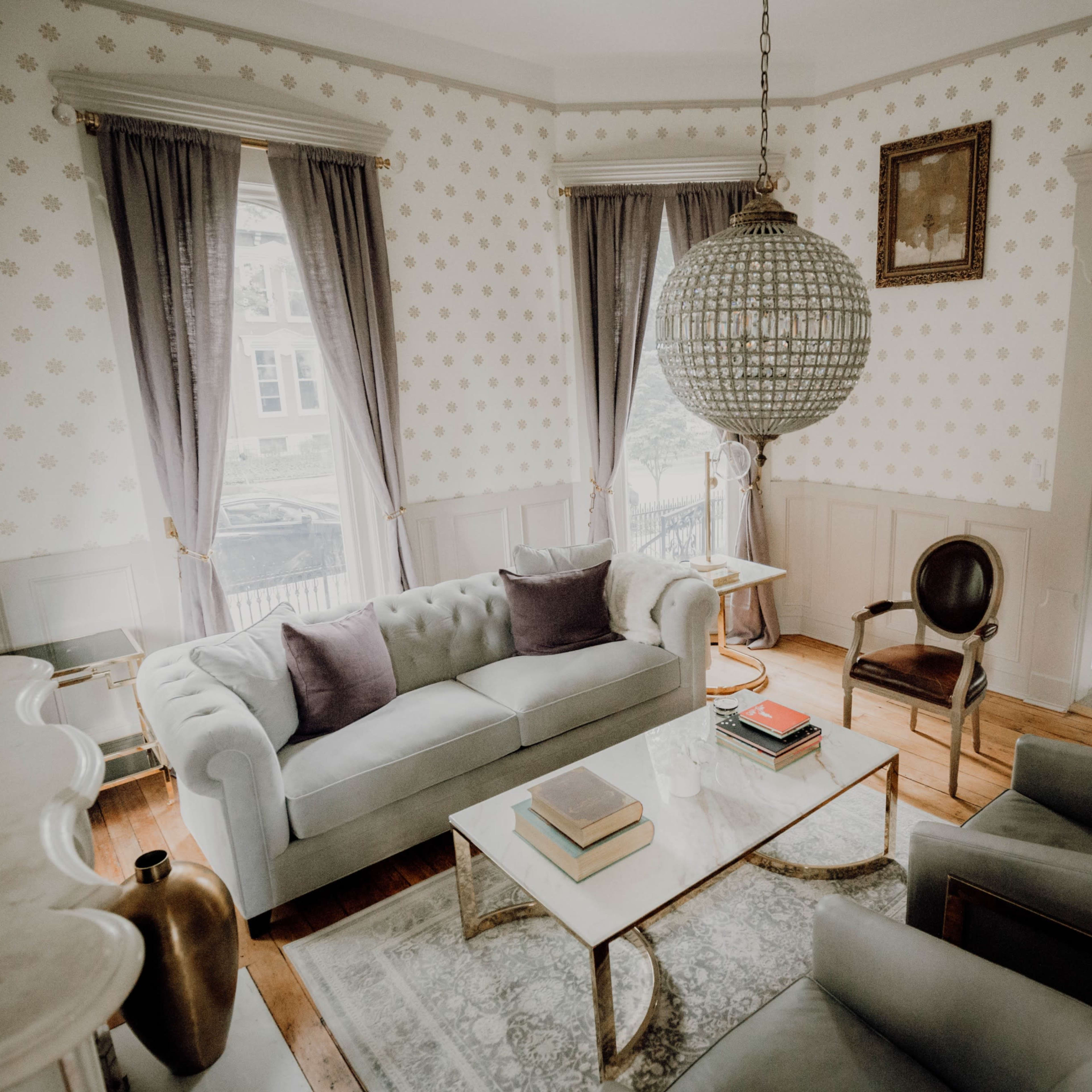 The room features a light-colored sofa with two dark cushions, a glass coffee table, and a large pendant light, with large windows dressed in curtains and a hardwood floor.