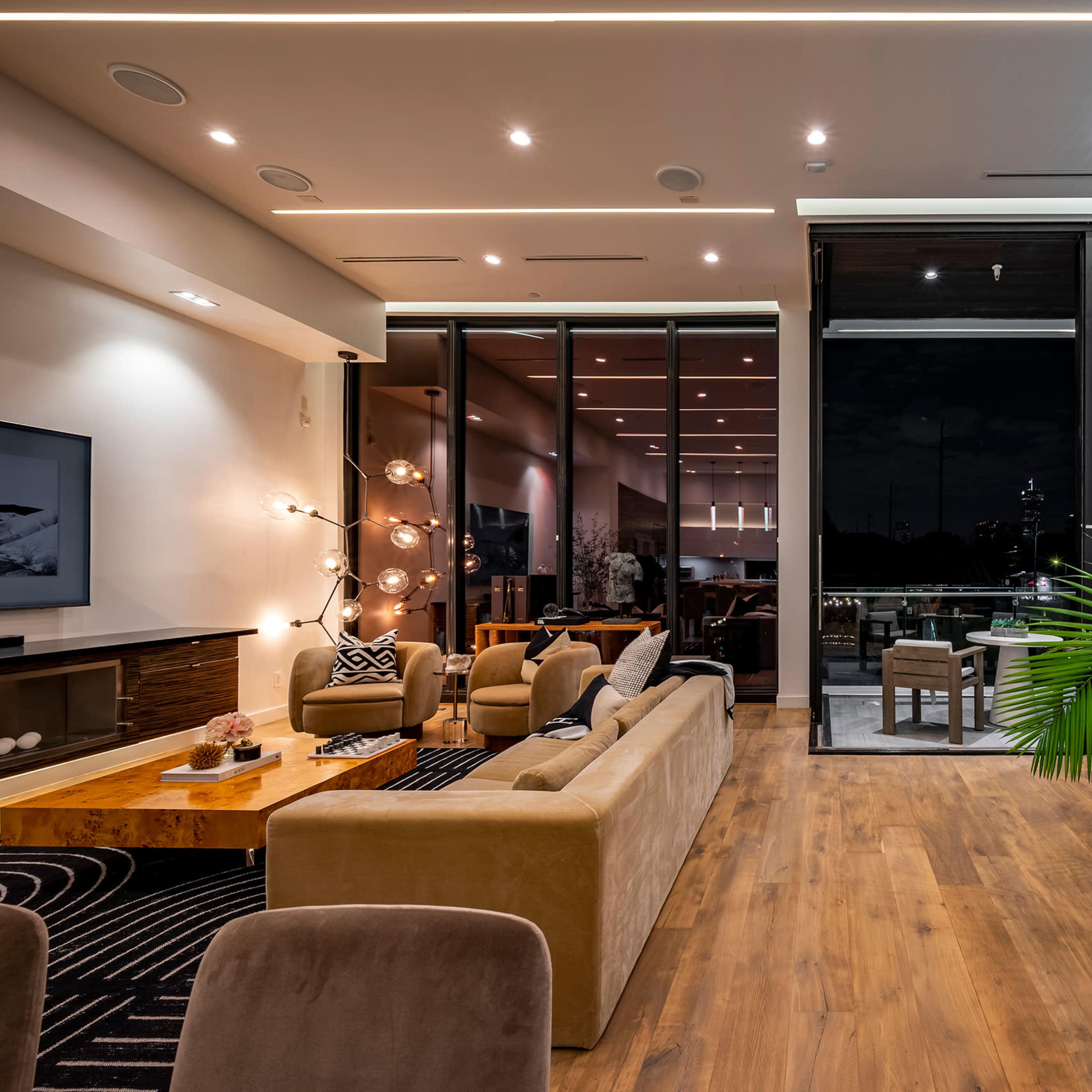 A modern living room with large windows, stylish furniture, and a decorative plant, all set against a backdrop of evening lighting.