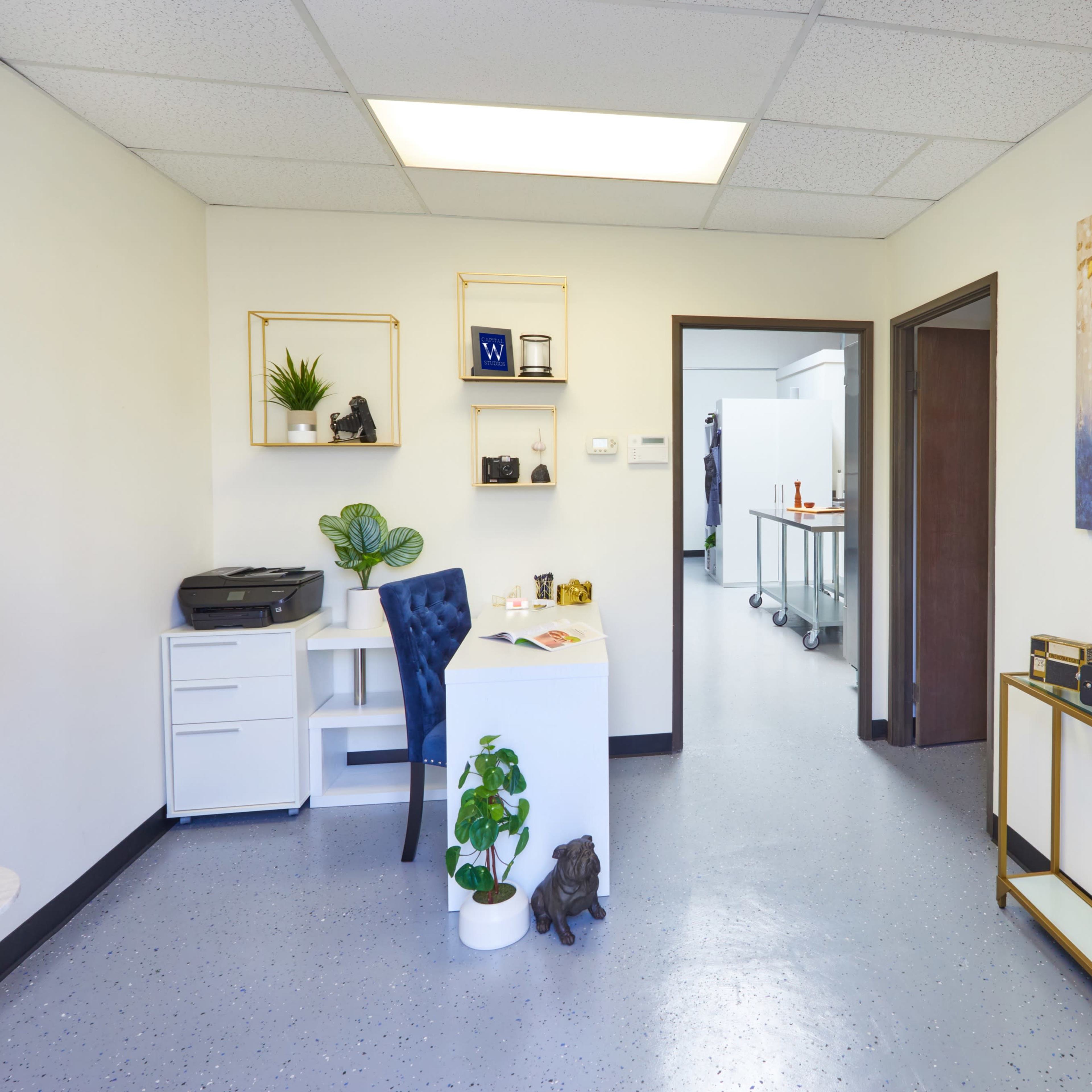 A small office space with a desk, a printer, a plant, and a shelf, leading to a doorway into another room.