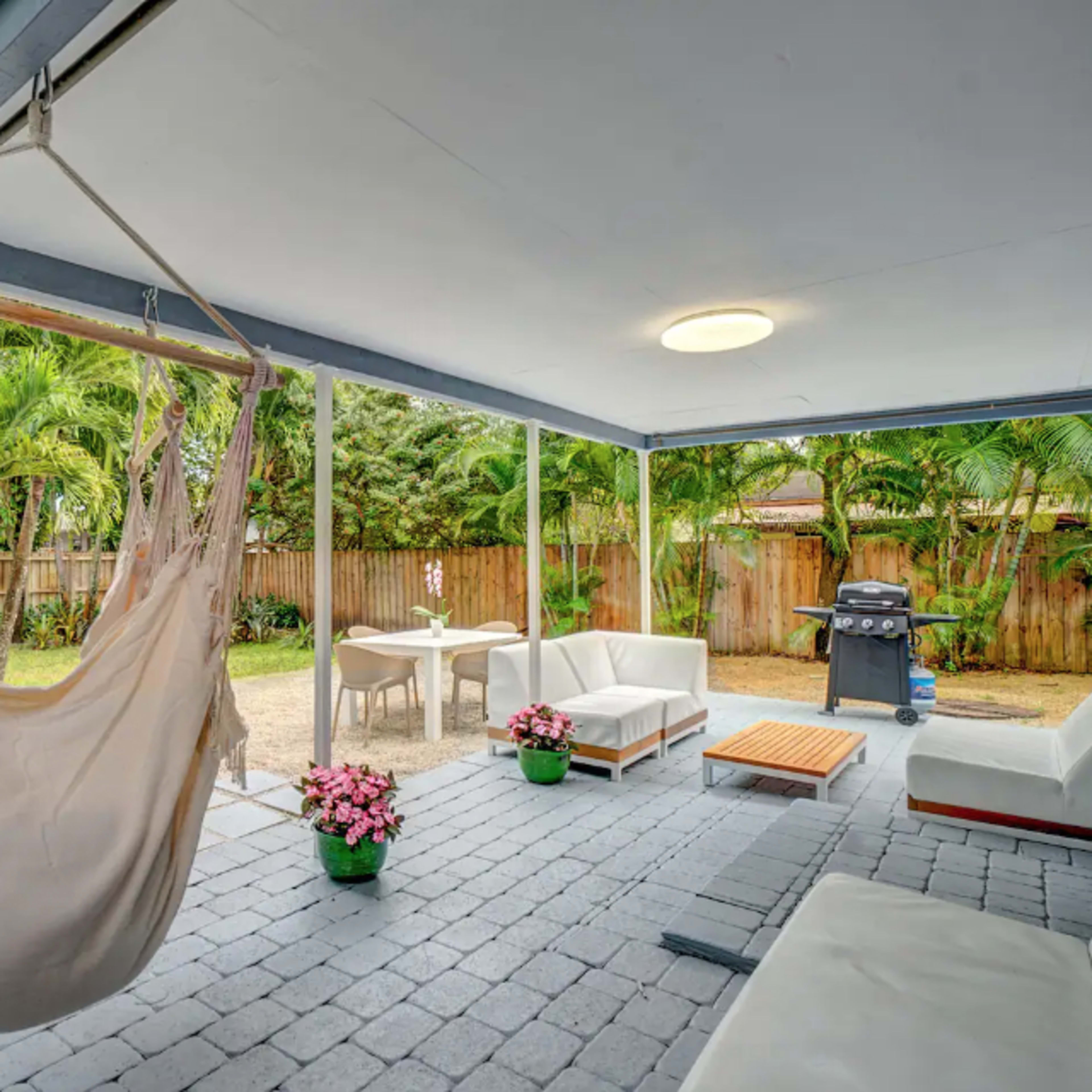 A covered patio with a hammock, seating area, a small table, and a barbecue grill, surrounded by greenery.