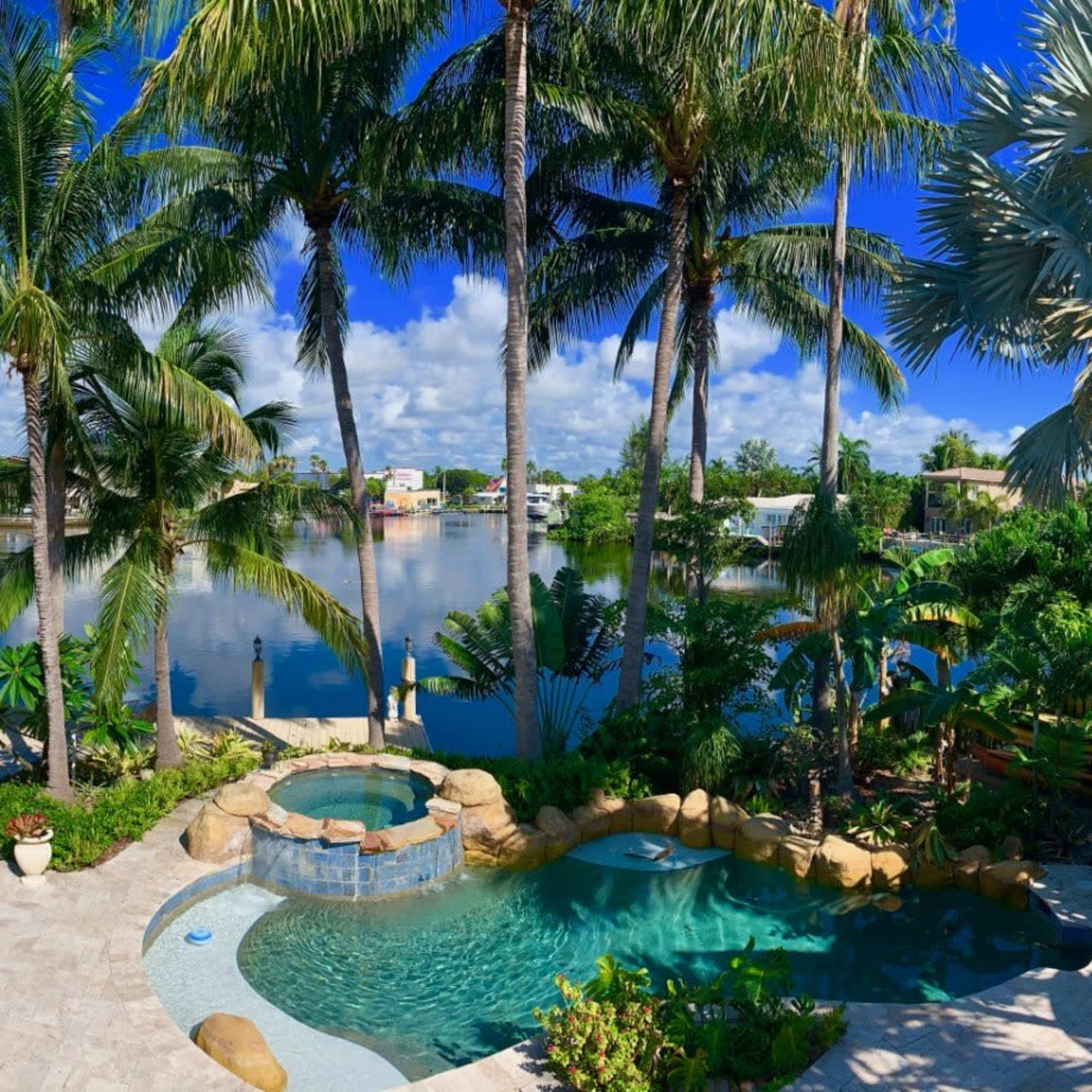 A landscaped backyard features a pool and spa area surrounded by palm trees and a waterfront view.