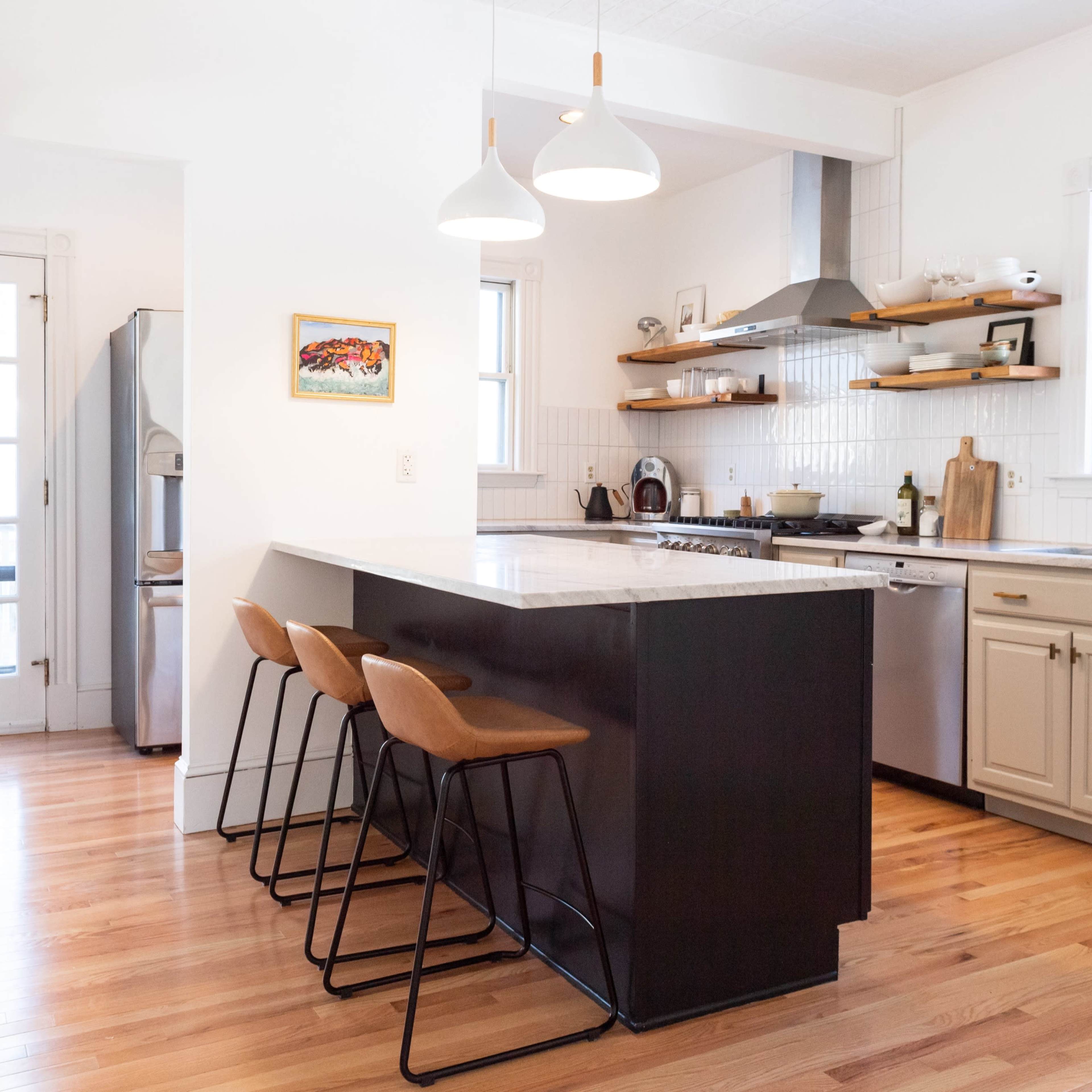 A modern kitchen with a central island and three bar stools, featuring white cabinetry, stainless steel appliances, and wooden floors.