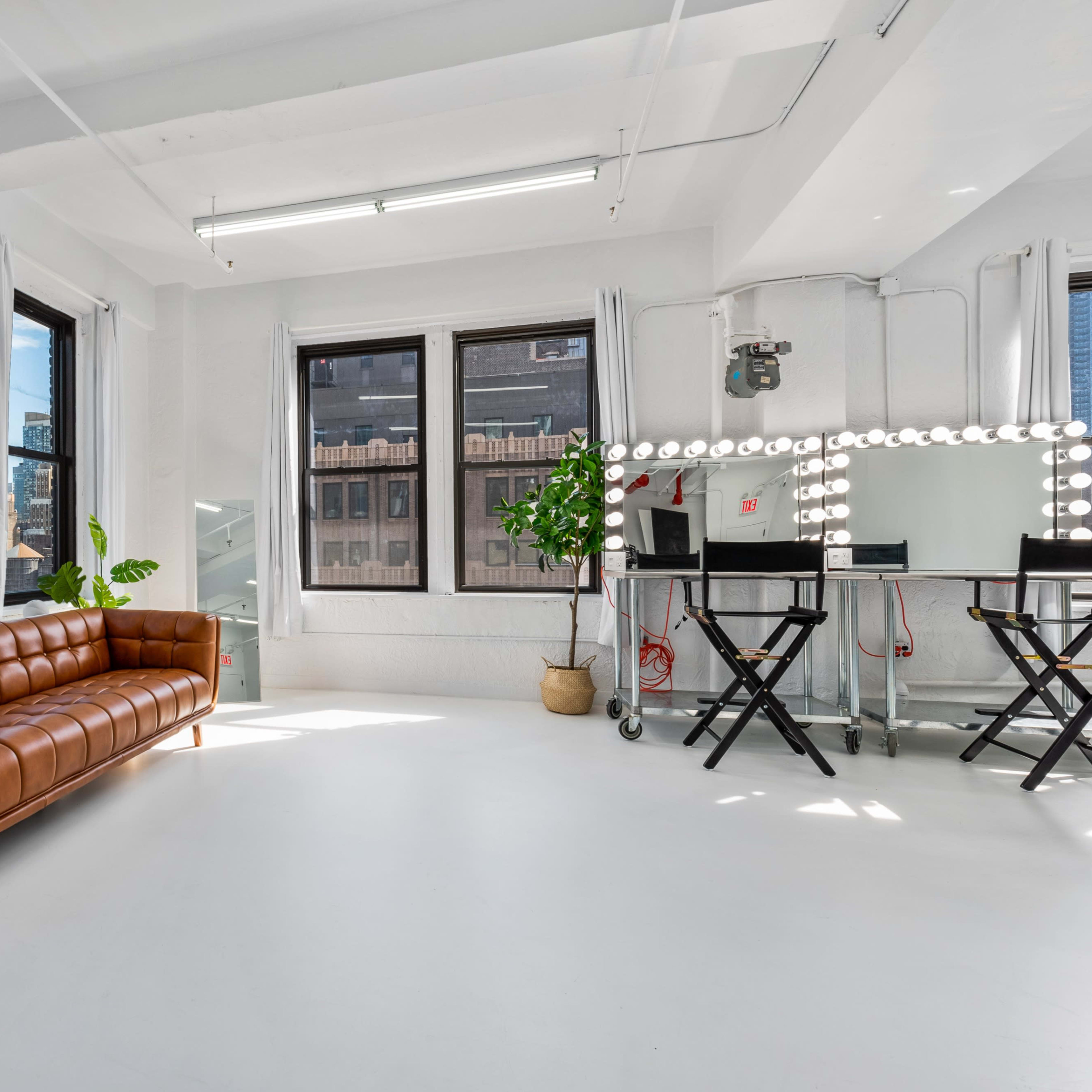 A bright, minimalist studio features a brown leather couch, a vanity with lights, and large windows showcasing the Empire State Building in the background.