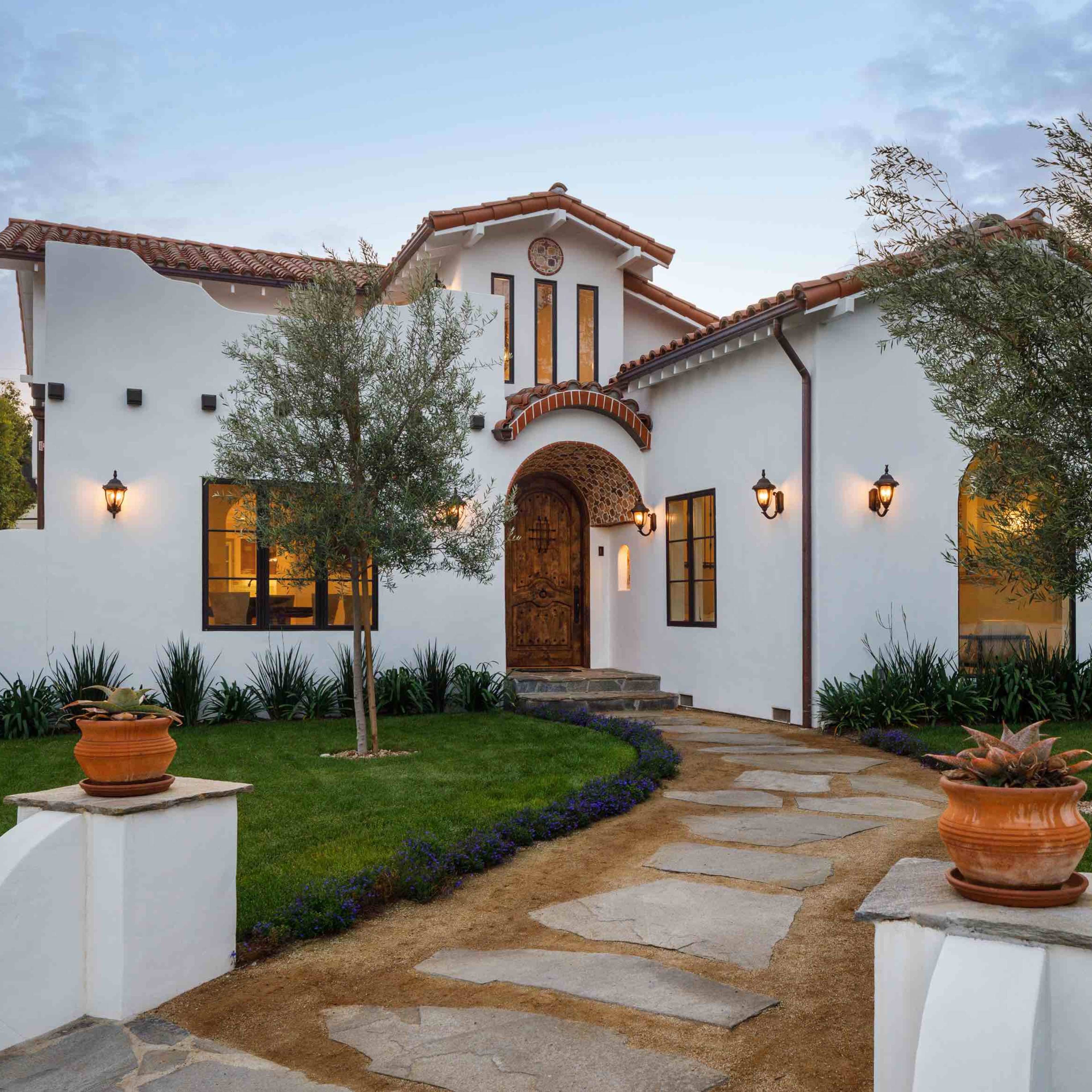 A white stucco house with a tile roof, surrounded by manicured landscaping and a stone walkway leading to the front entrance.