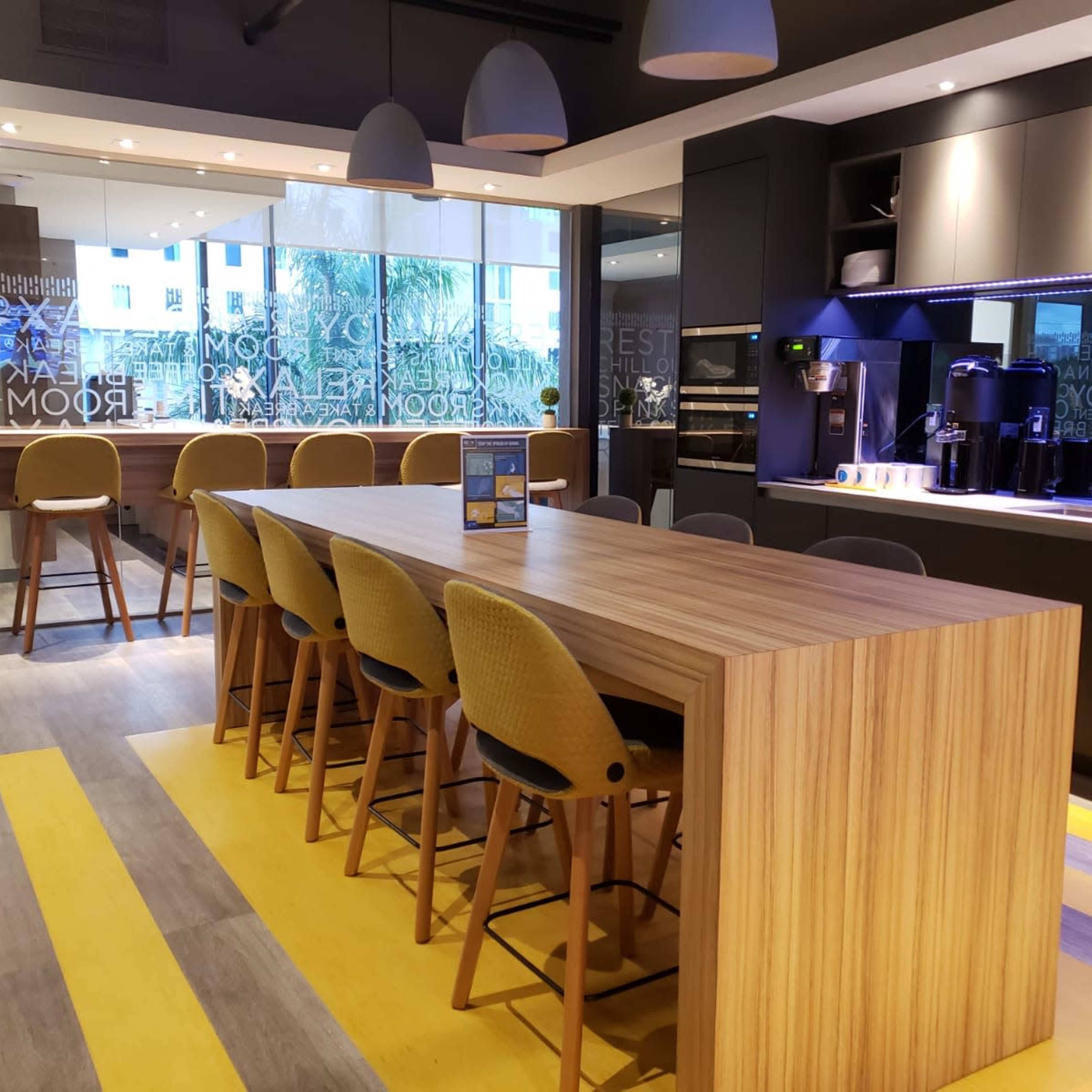 A modern kitchen space featuring a long wooden table surrounded by high-backed stools, with appliances and a bar area in the background.