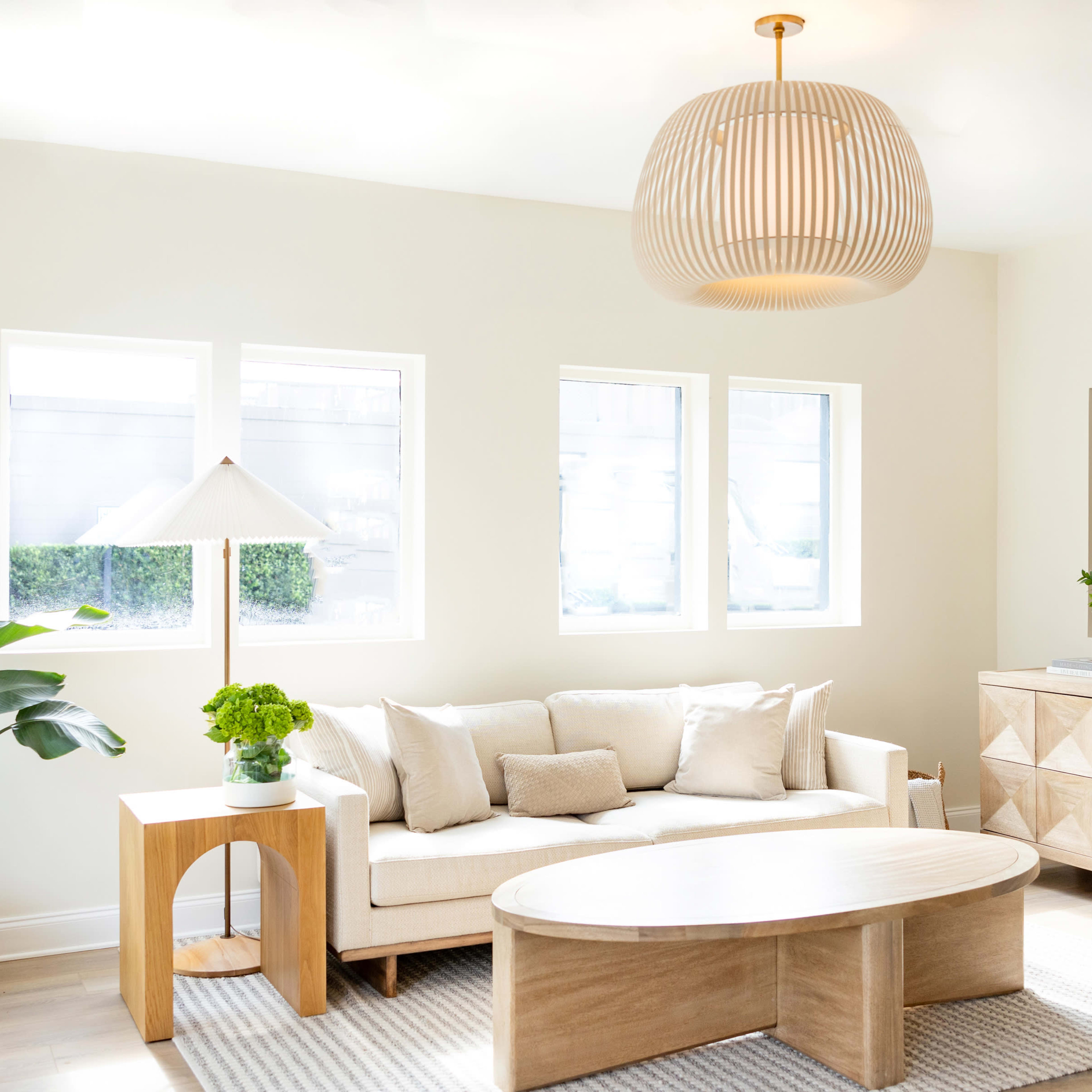 A bright living room featuring a beige sofa, a round coffee table, a decorative plant, and a wooden sideboard under a woven pendant light.