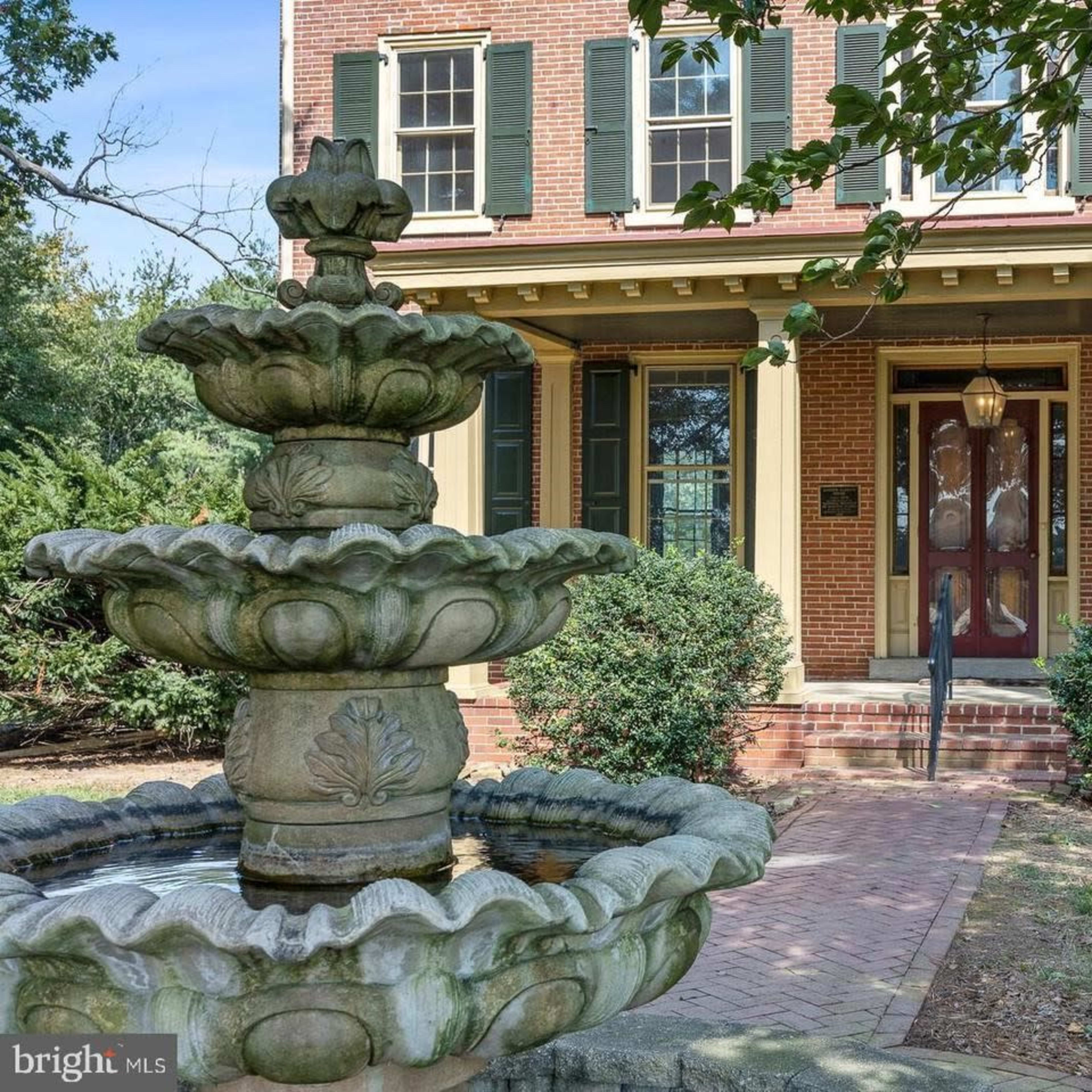 A brick house features a front porch with green shutters and a three-tiered fountain in the foreground.