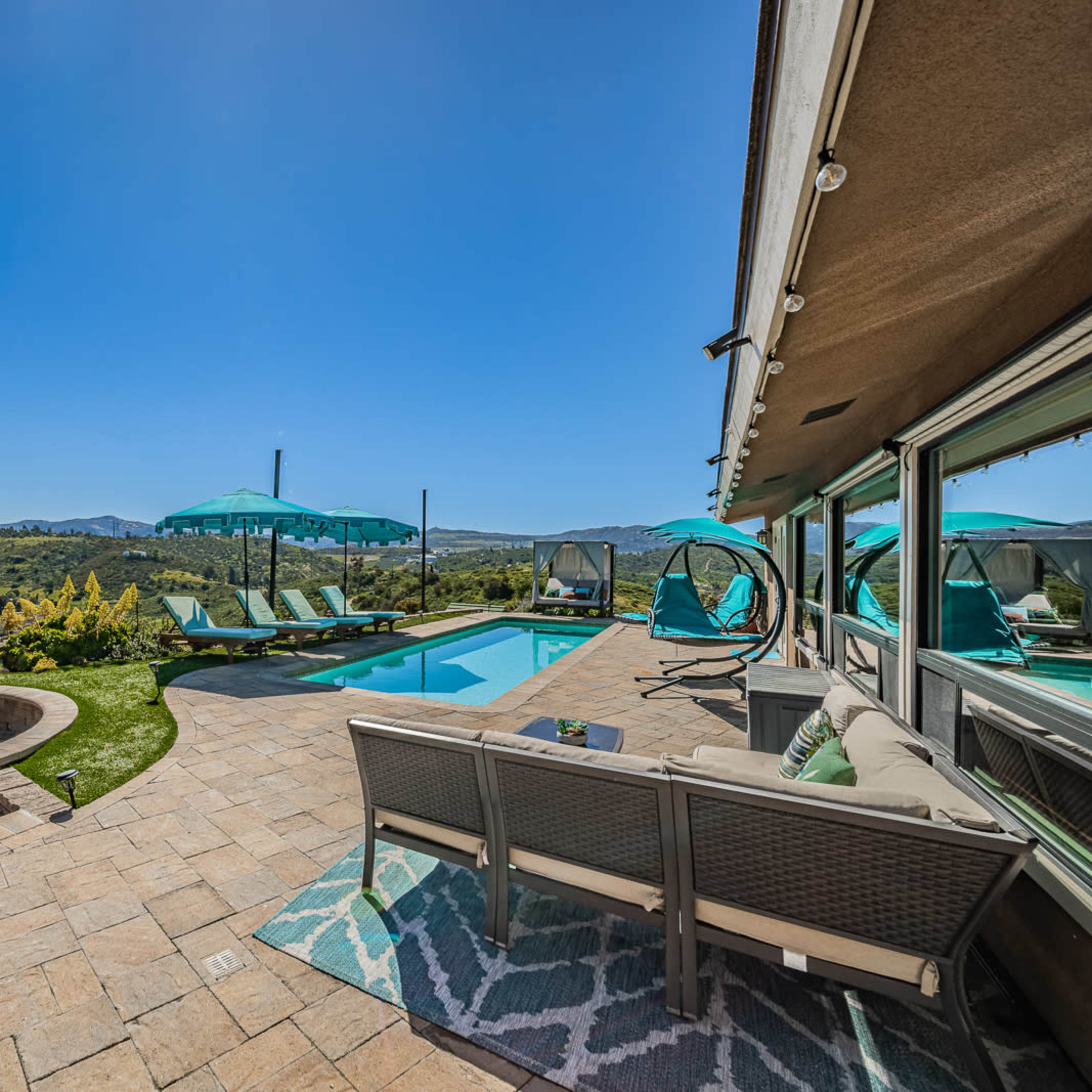 A backyard pool area with lounge chairs, a hot tub, and scenic hills in the background under a clear blue sky.