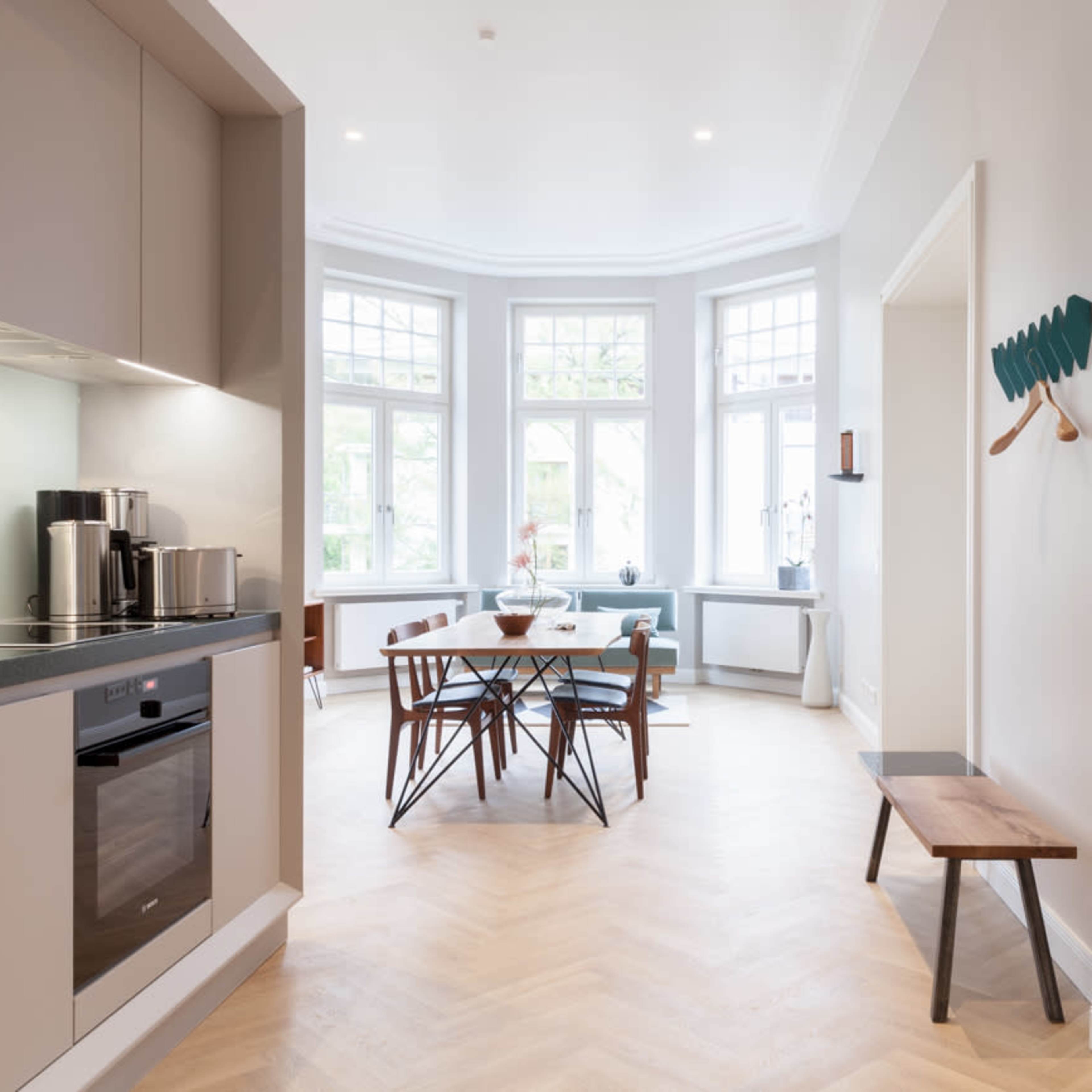 A bright, modern kitchen opens into a dining area with large windows and a wooden table surrounded by chairs.