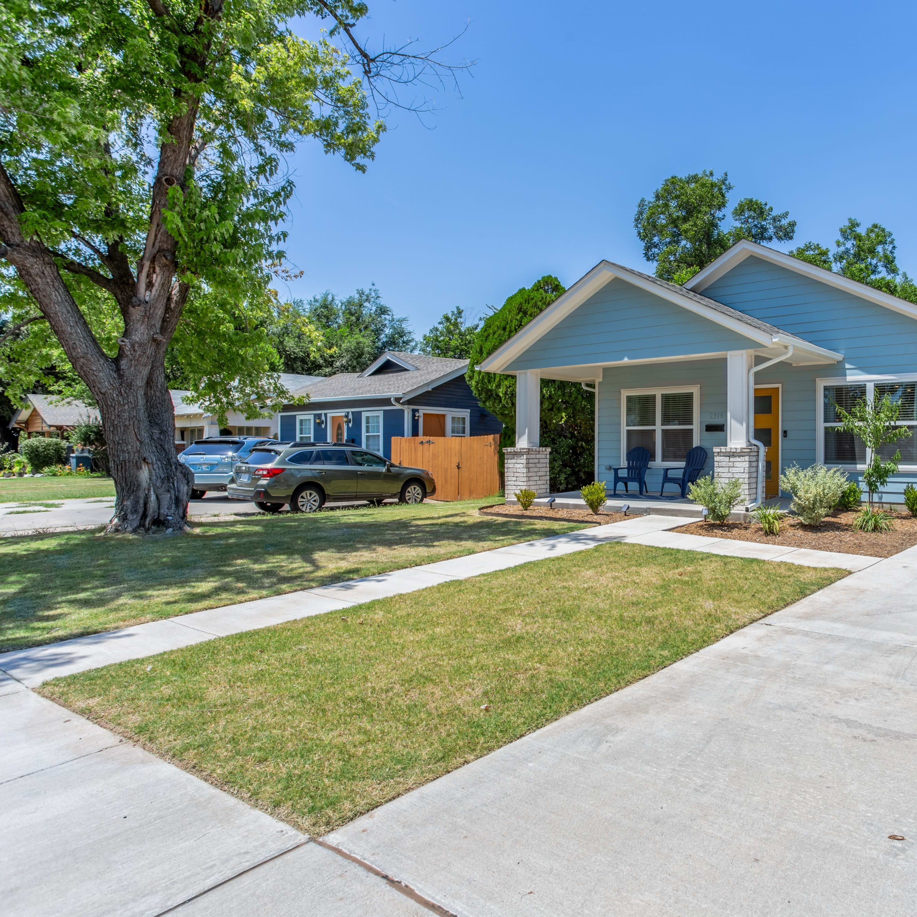 A light blue single-story house with a front porch is situated on a landscaped lawn, flanked by trees on a sunny day.